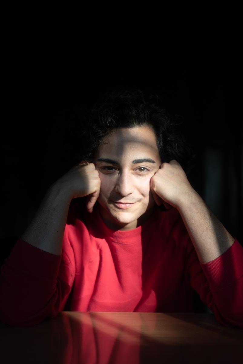 A person in a bright red shirt sits at a table with their elbows resting on the surface and their hands gently framing their face. Soft sunlight creates a diagonal band of light across his eyes, highlighting his warm, subtle smile. Their dark, curly hair blends into the deep shadow behind him.