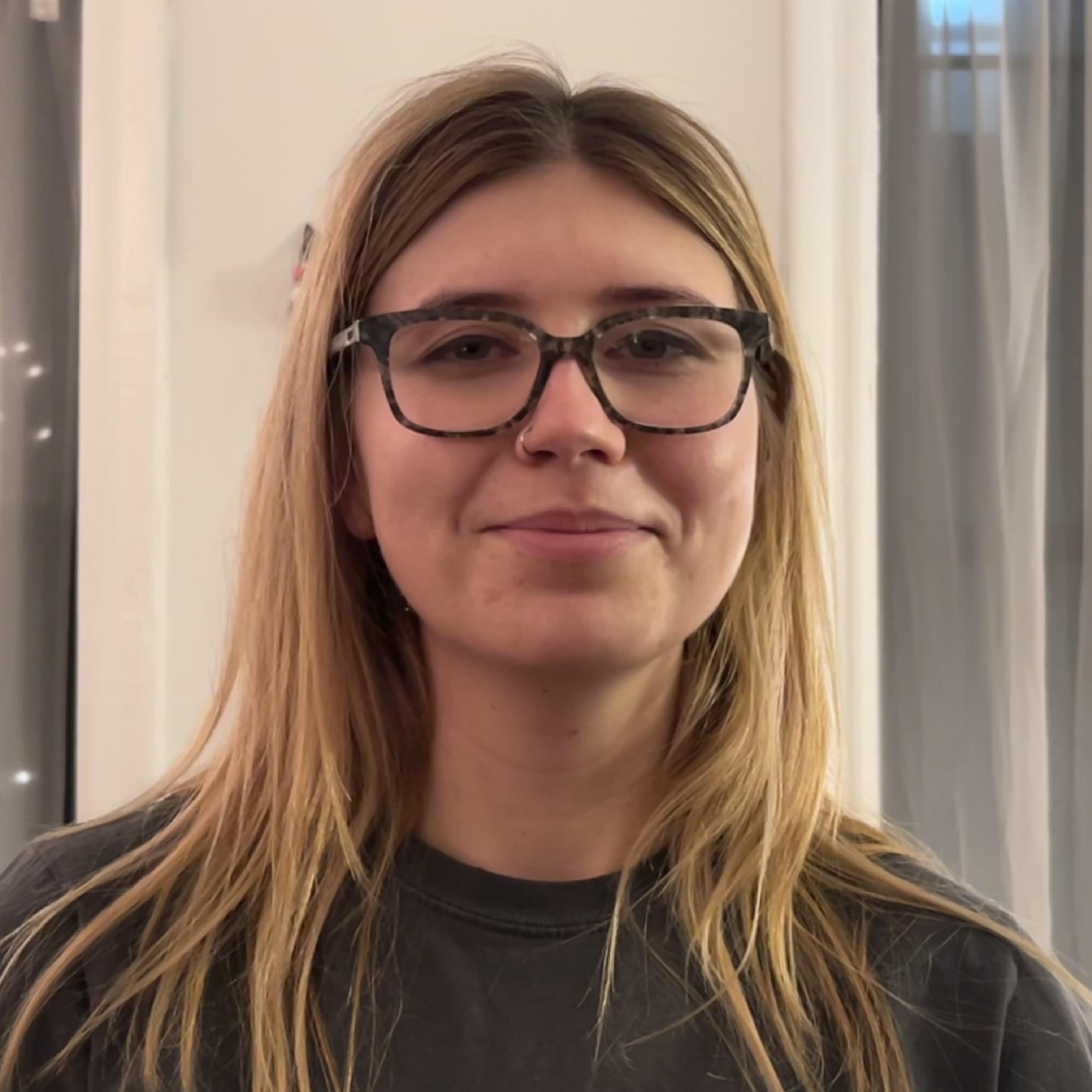 A white woman with blonde hair, glasses, and a gray t-shirt. She is standing in front of a white wall with sheer curtains on either side.