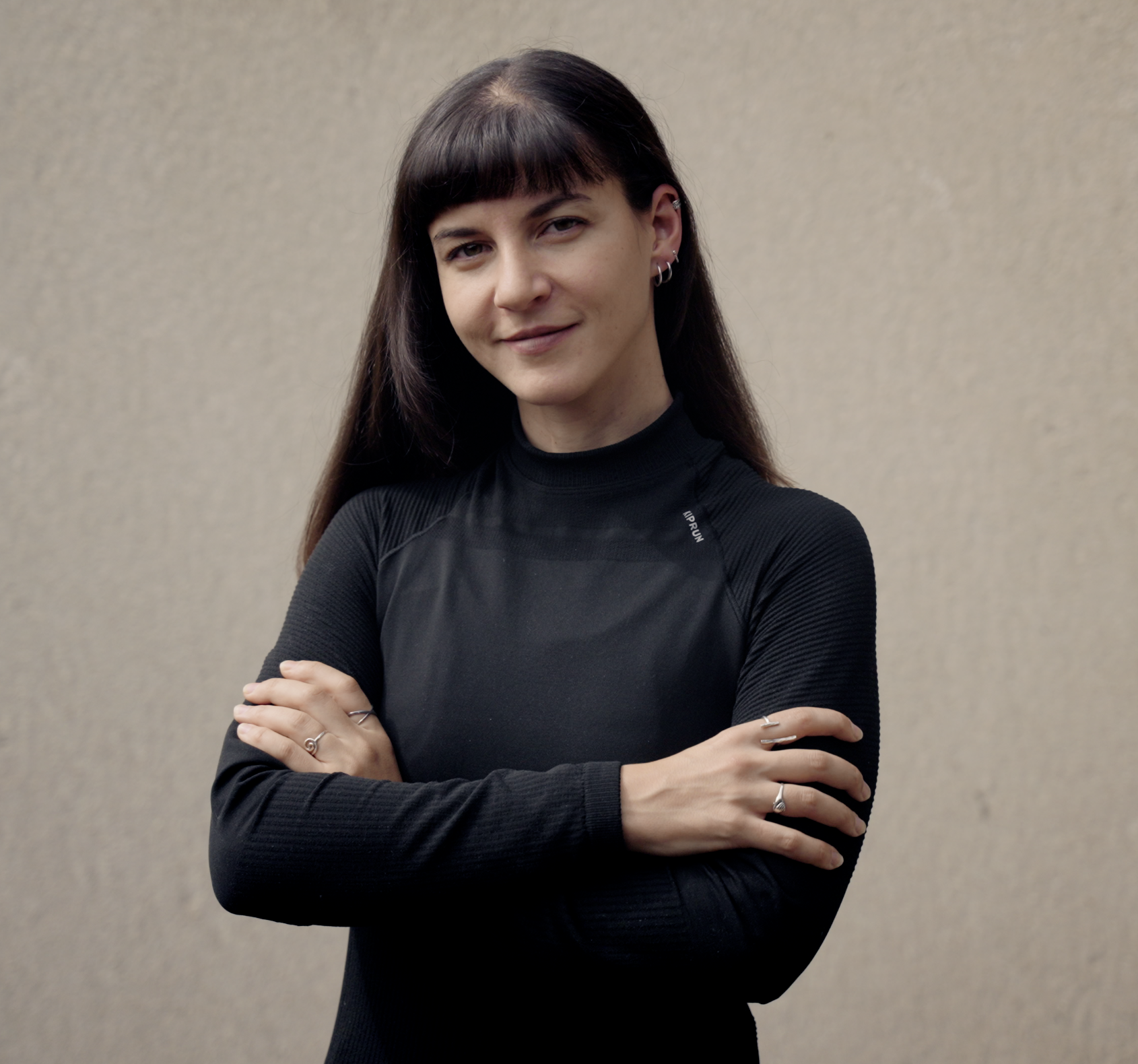 A woman stands with her arms crossed in front of a beige wall and looks into the camera.