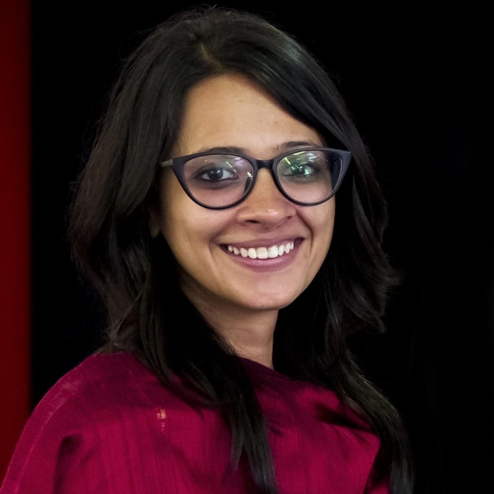Headshot of Shirley Abraham- she has black hair and is wearing black-rimmed spectacles and a red scarf.