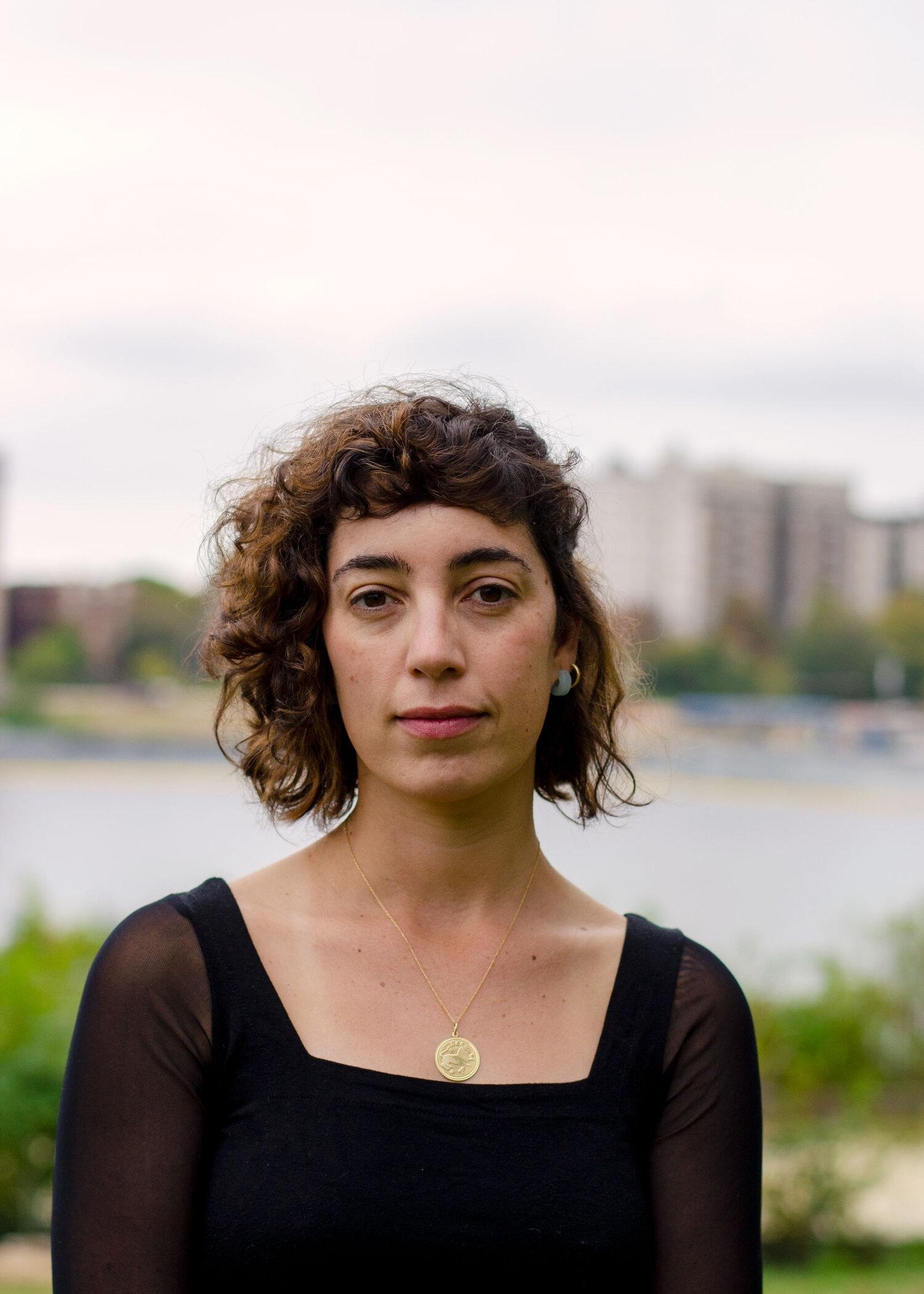 A warm color portrait of filmmaker Jonna McKone standing outdoors in daylight and looking at the camera. She has short curly brown hair and is wearing a black dress, a slender chain around with a round pendant. The background is out of focus with some greenery, a small lake, and a few buildings.