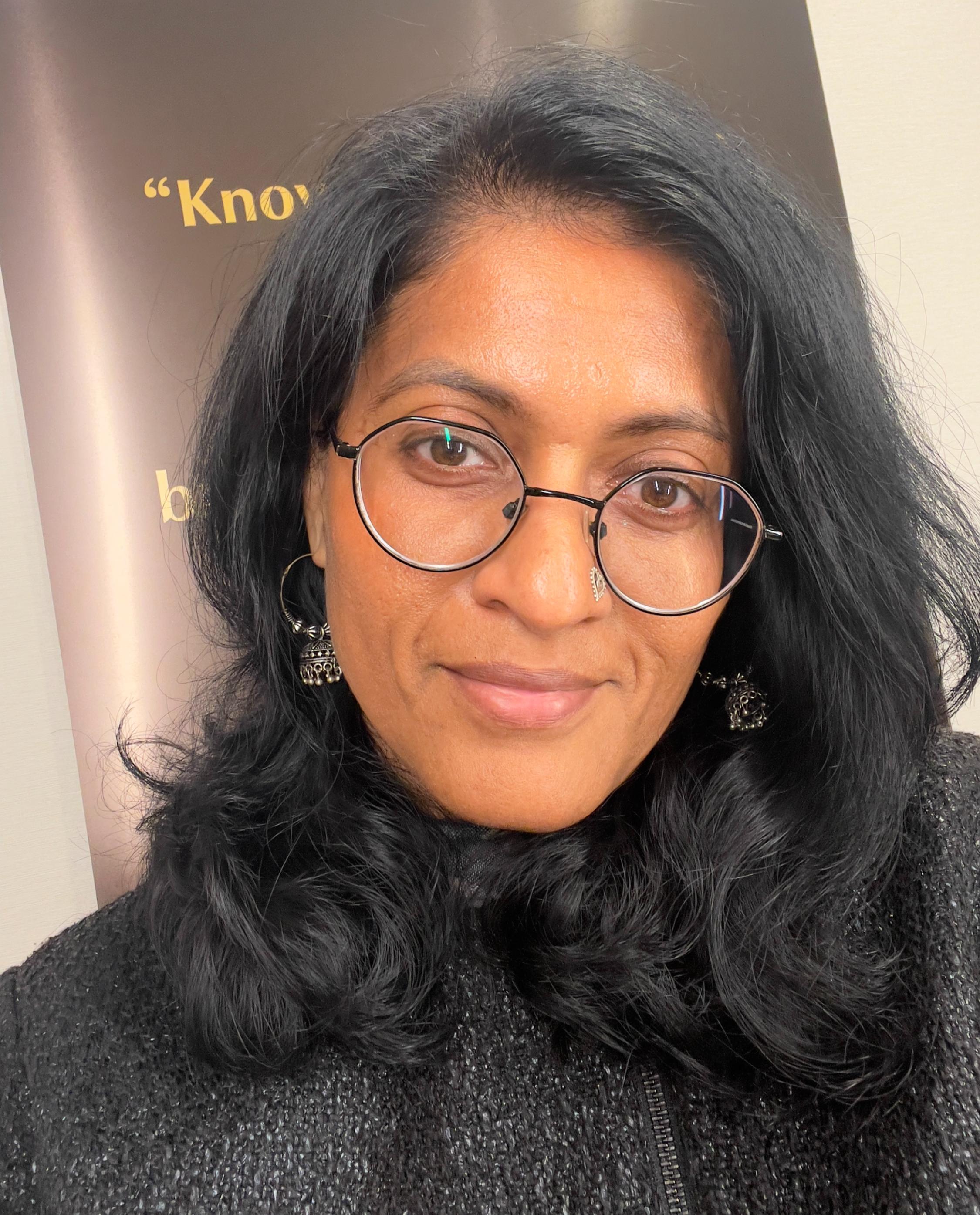 A close-up selfie portrait of a South Asian woman with dark wavy hair, wearing round black-rimmed glasses, silver jhumka earrings, a nose pin, and a slight, warm smile.
