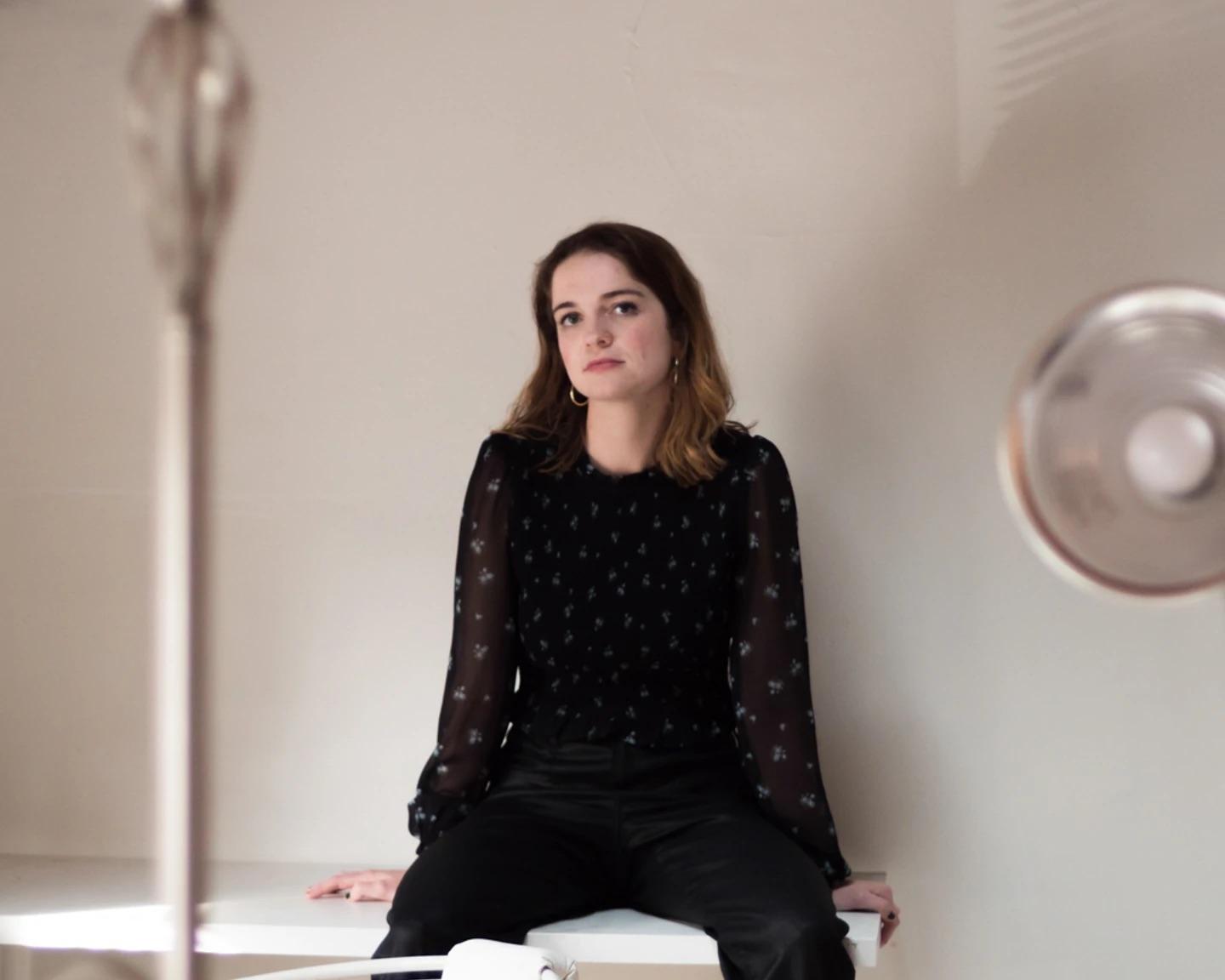 A woman with shoulder-length brown hair wearing a black blouse and pants sits on a white surface in a neutral-toned room, looking directly at the camera.