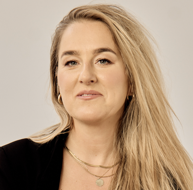 A woman with long blonde hair smiles softly at the camera. She wears gold hoop earrings, layered gold necklaces, and a black top, against a light neutral background.