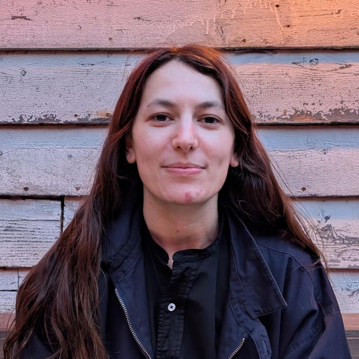 A white woman with long brown hair and a blue jacket sits in front of a wooden shingled wall. She is slightly smiling.