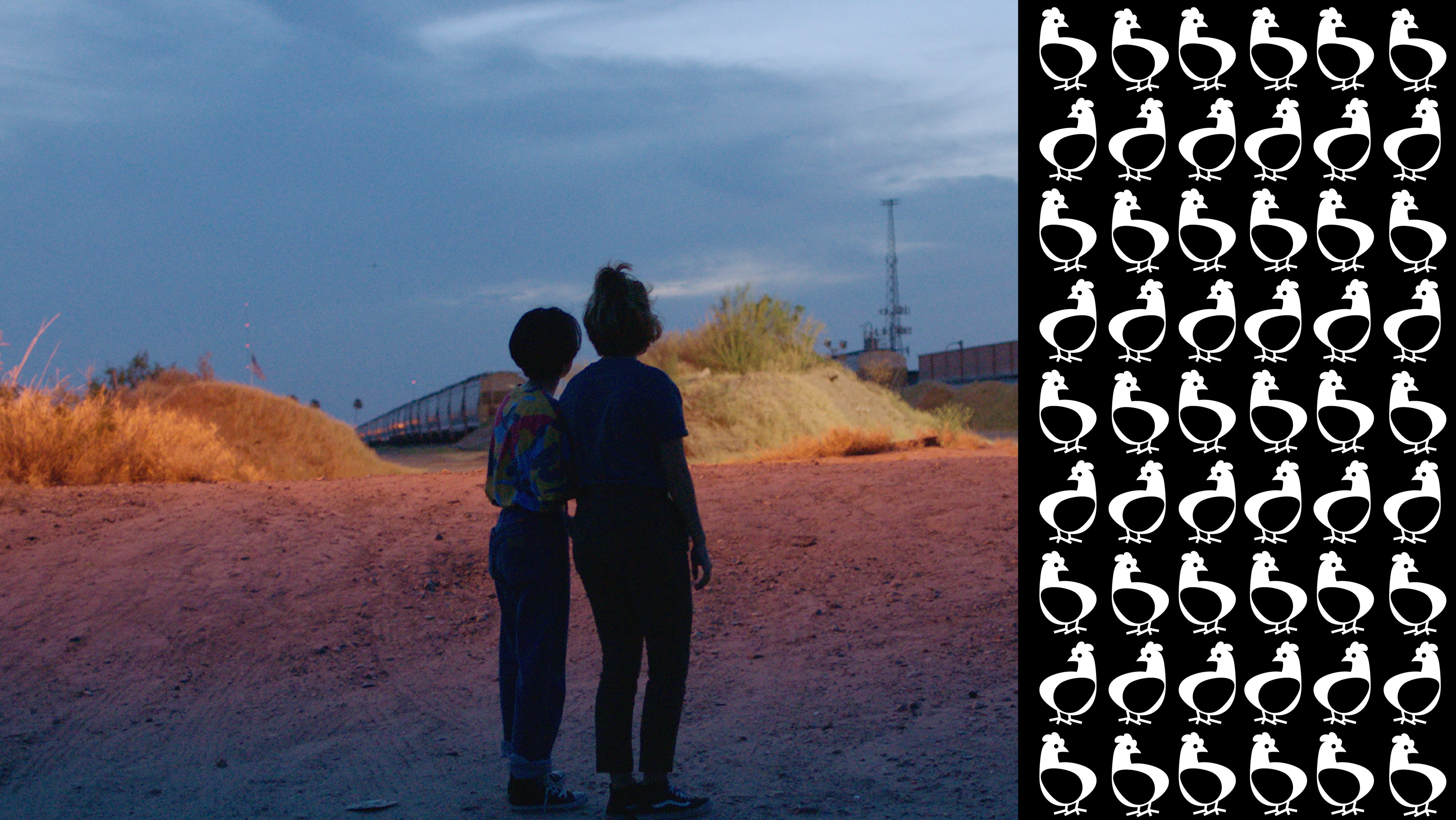 A color still from the film Hummingbirds, in which two people are standing in the foreground on a sandy outdoor landscape with a train approaching from their left.
