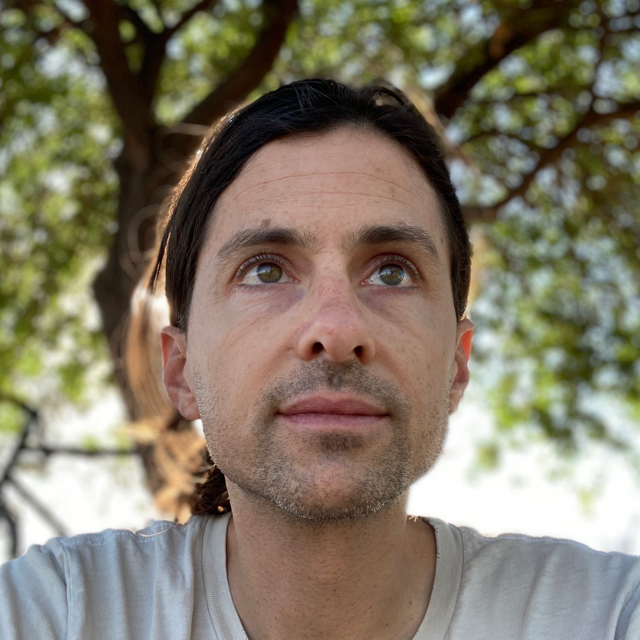 Portrait of Nick Kelso. He wears a shirt and looks away from the camera, in the background there is a tree. The portrait is taken in a low angle.