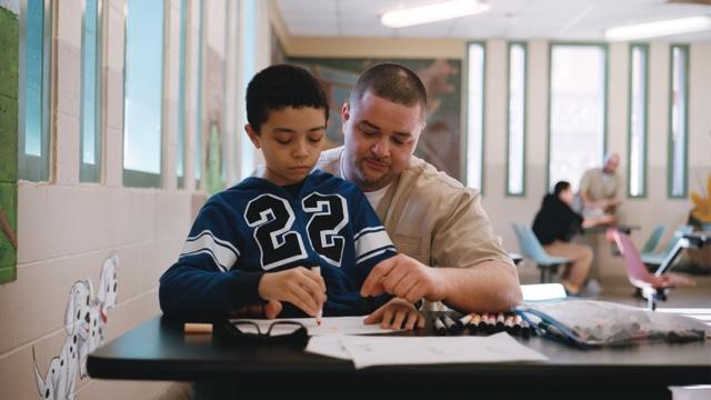 An incarcerated father helps his son with a coloring page at visitation.