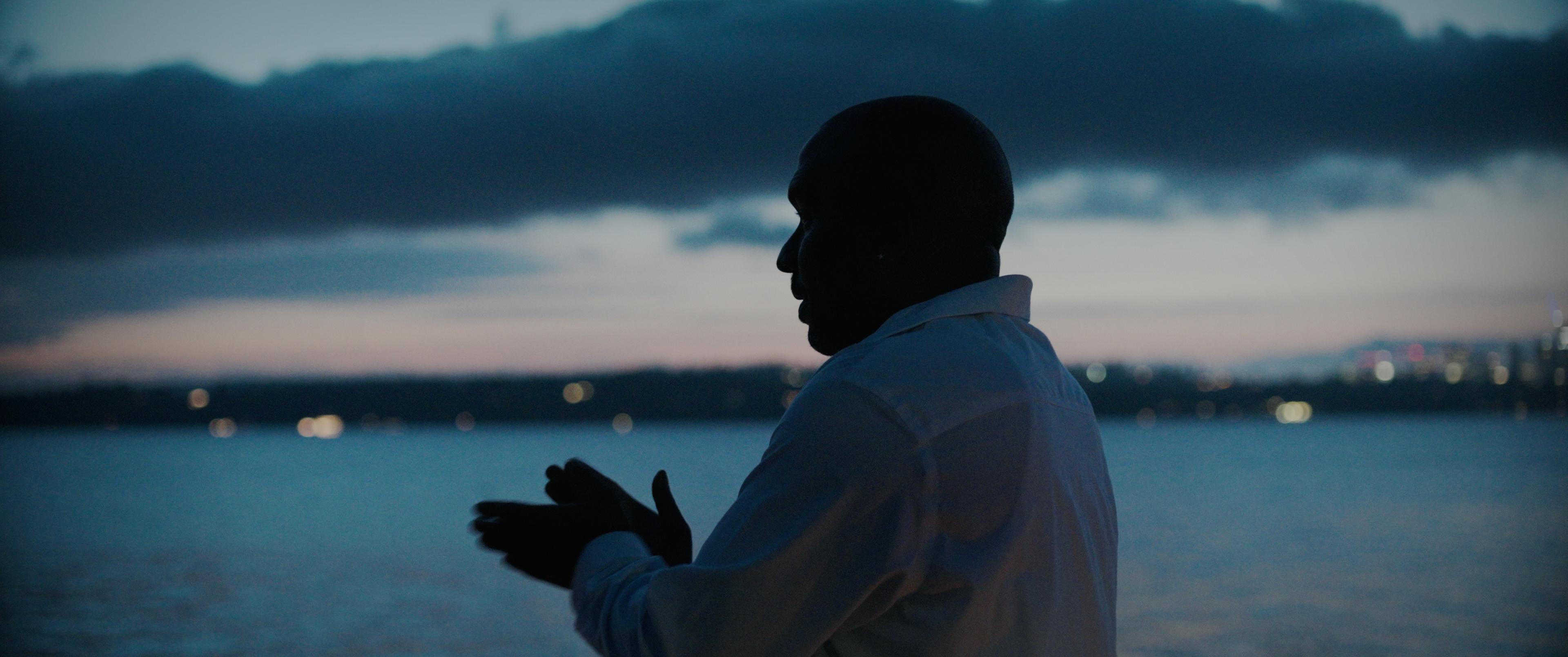 A man stands on a beach at dusk. He is backlit and presses his hands together.