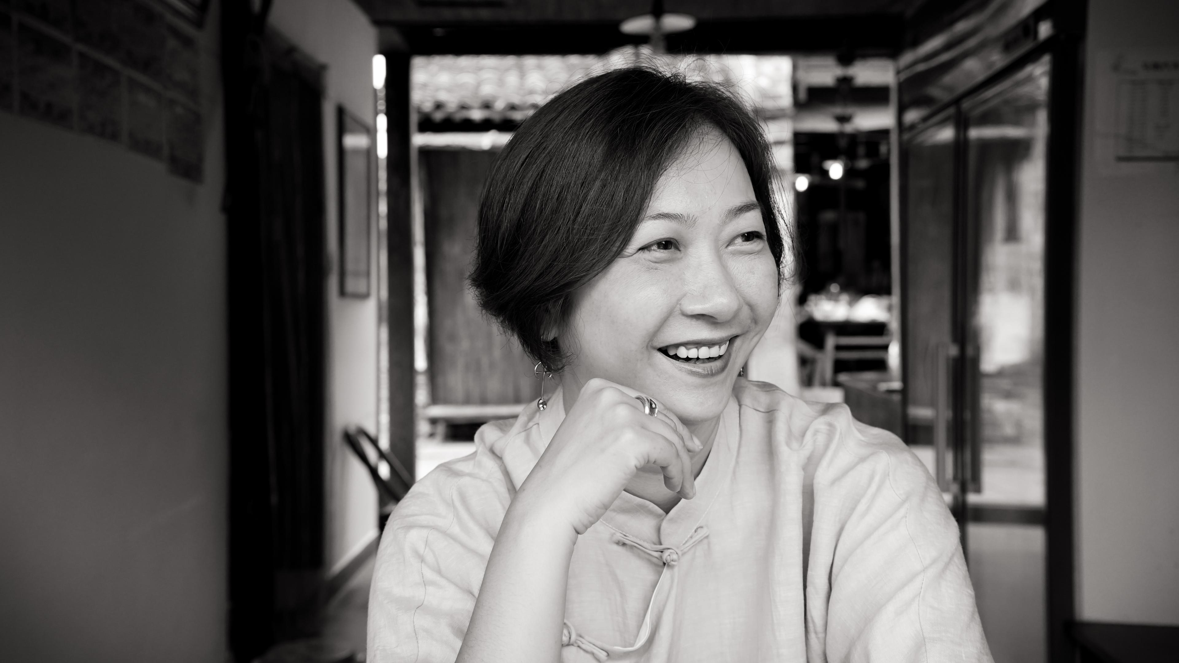 Zhao Qing looking to her side and smiling. She has short dark hair, and is wearing a light-colored blouse. Black and white portrait, with out of focus background.