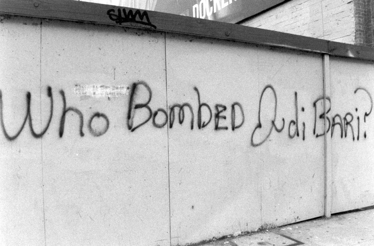 A black and white photo of a graffitied wall that reads "Who bombed Judy Bari?"
