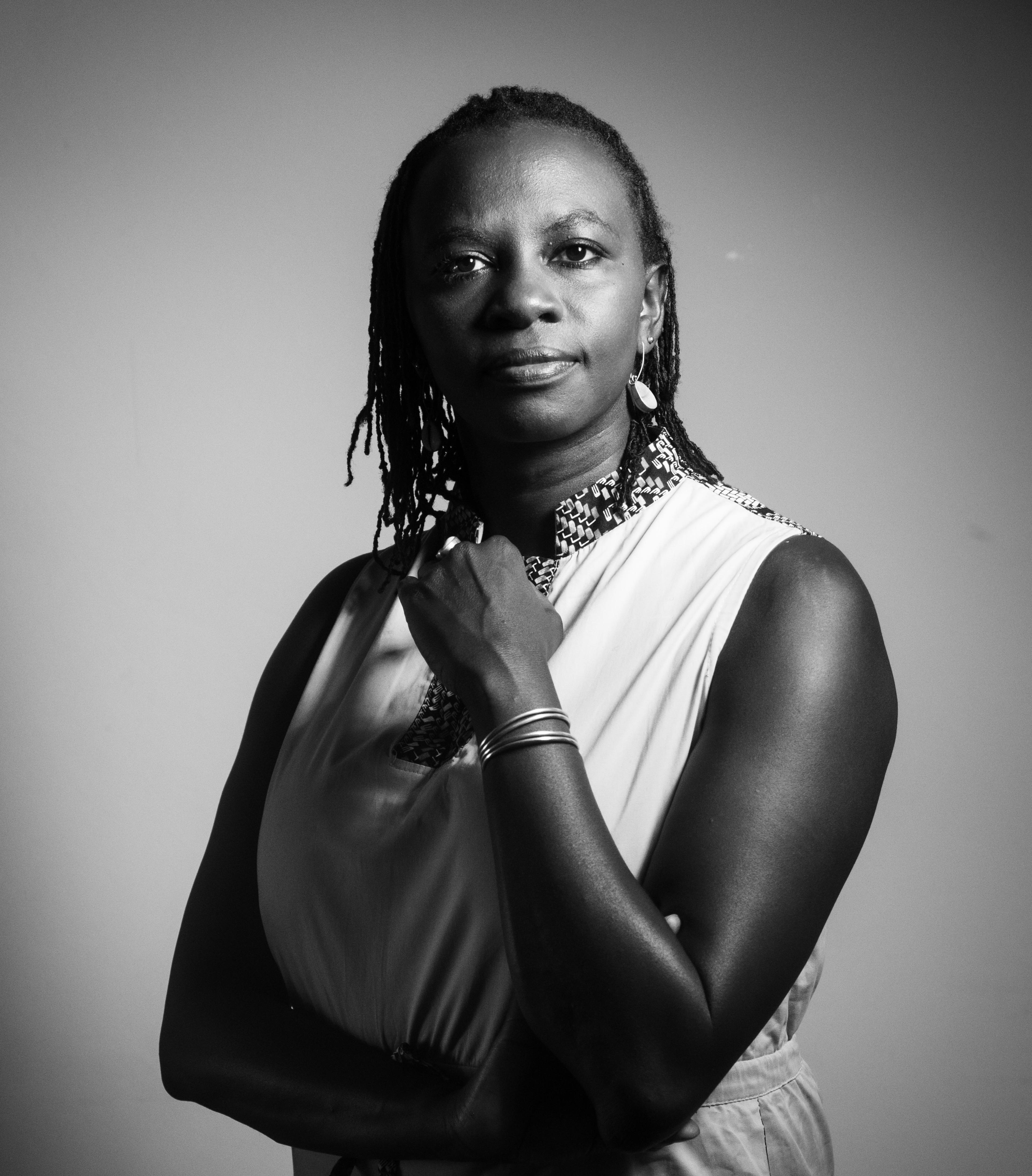 A dark-skinned woman standing in dim, shadowy lighting, looking straight ahead with one arm folded and the other resting across her chest.