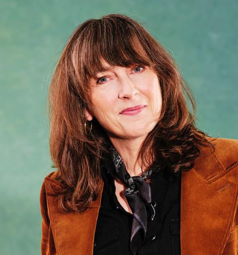 A woman with long, wavy brown hair and fringe poses against a blue background. She is wearing a rust-colored corduroy blazer over a black shirt and a dark patterned scarf tied loosely around her neck.