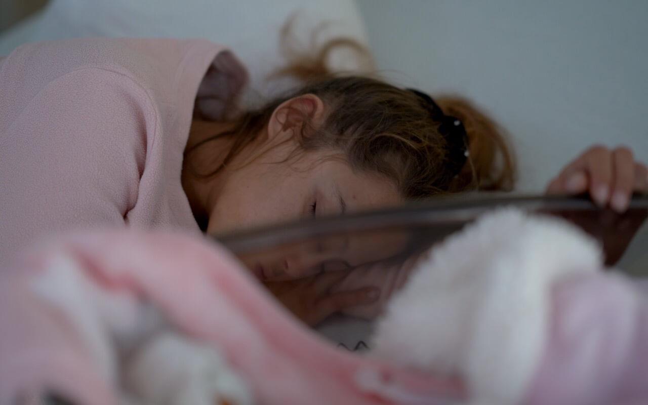 A little girl sleeps on her bed holding a mirror that shows her reflection.