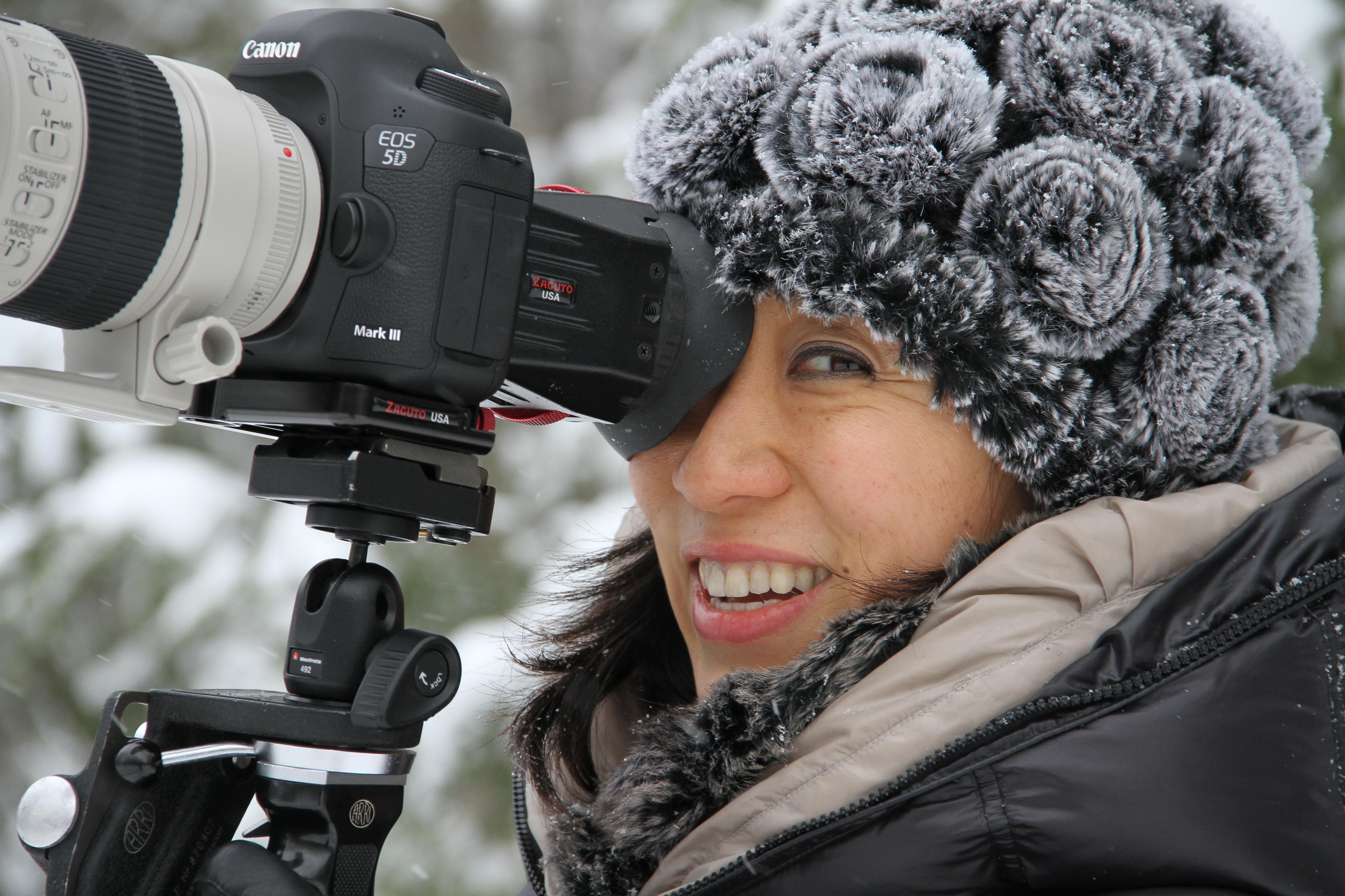 A woman looking through a camera eyepiece smiles at the camera.