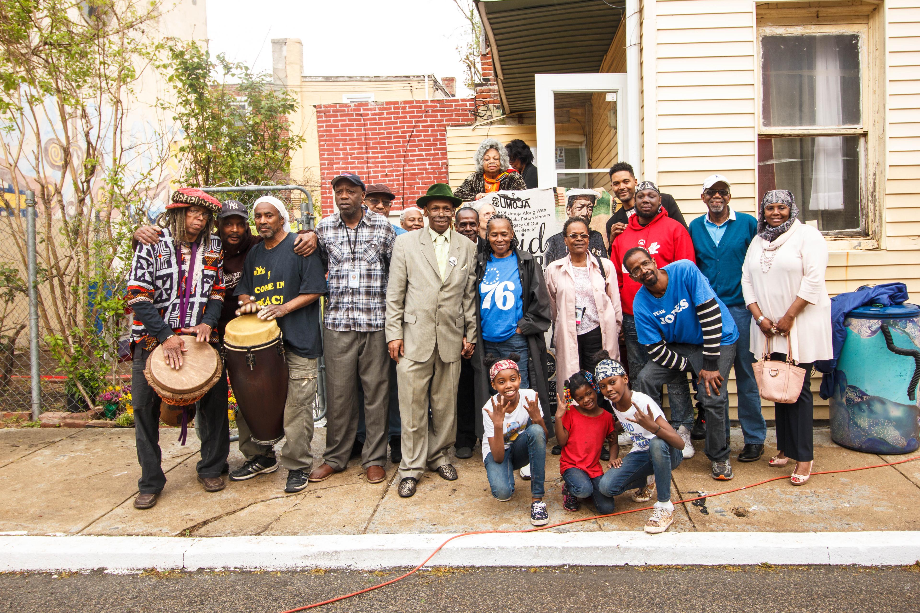 Still from Falaka Fattah and The House of Umoja. A big group of people standing by a house, some of which are holding drums, smile and look at the camera.