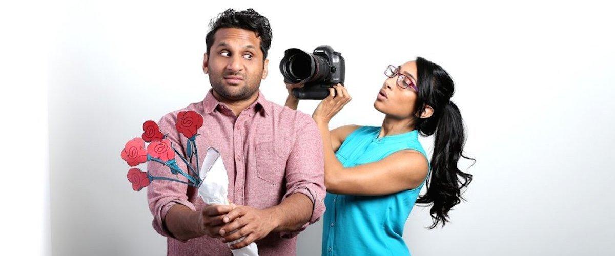 A man holds a bouquet of paper flower while a woman stands behind him and holds a camera up to his face.