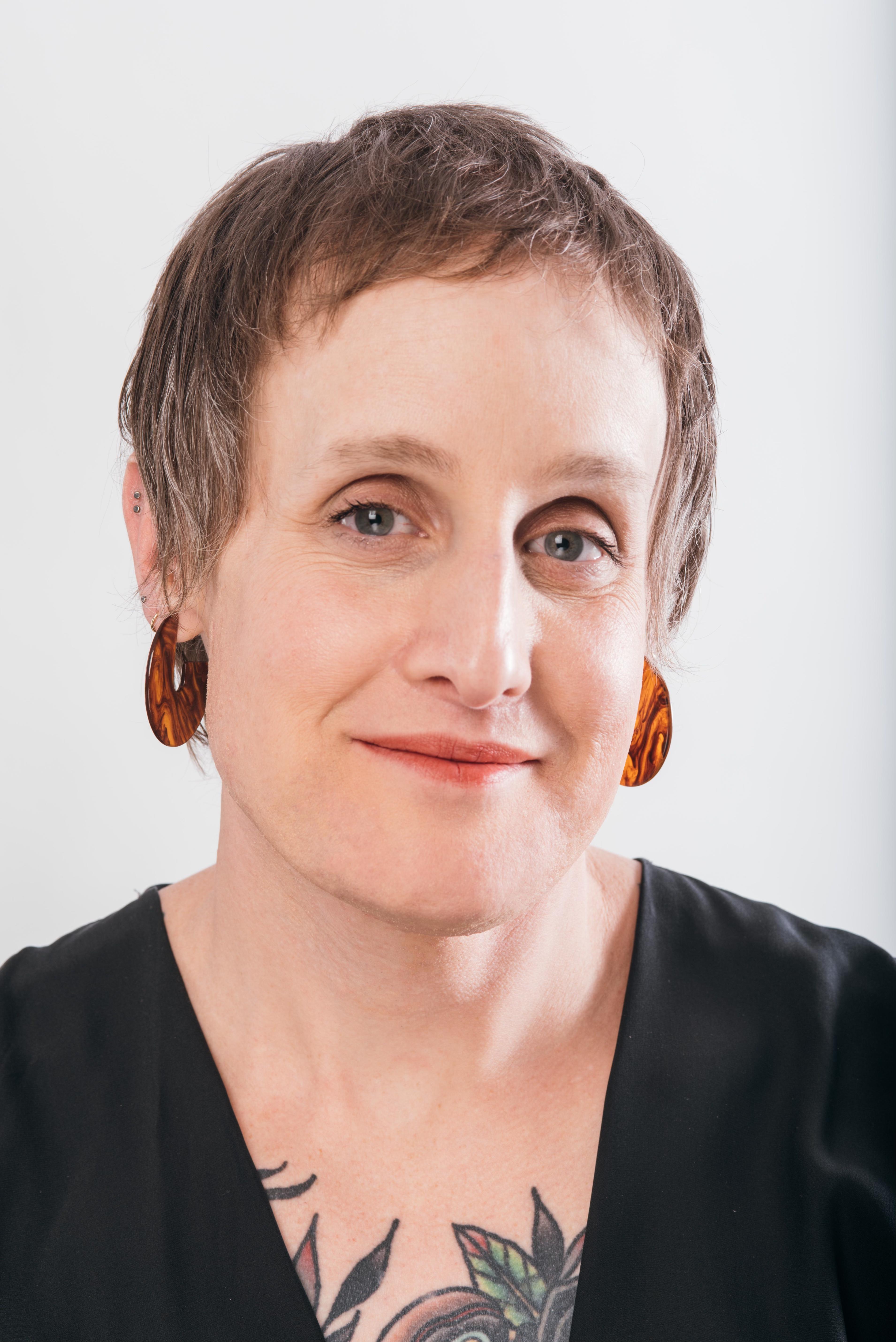 White woman with brown hair and blue eyes and tattoos wears a black dress and hoop earrings.