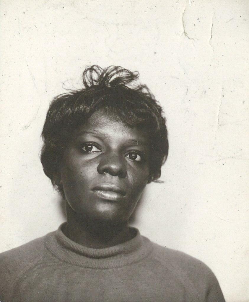 An archival photo of a woman wearing a turtleneck standing against a plastered wall.