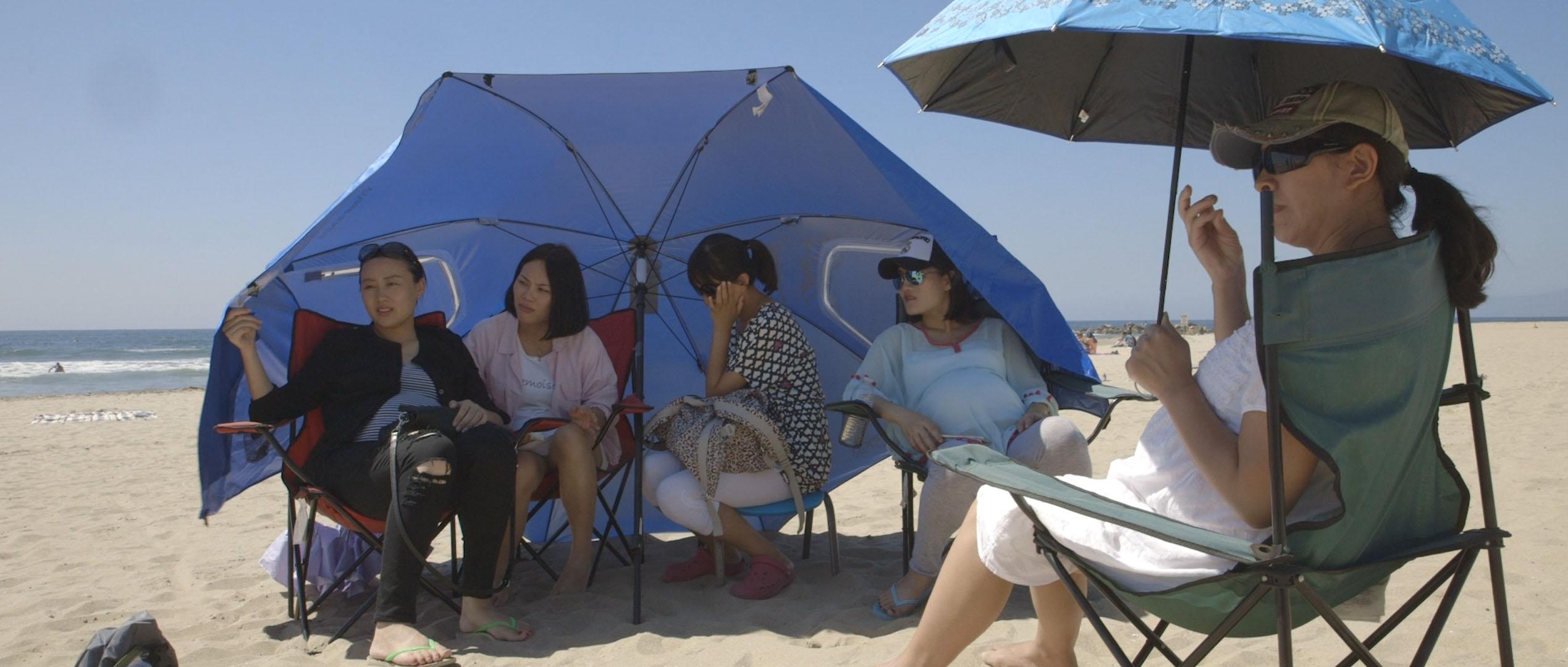On a beach, a group of women sit on beach chairs under an umbrella. In the foreground, another woman sits holding an umbrella. They all look off to the side.
