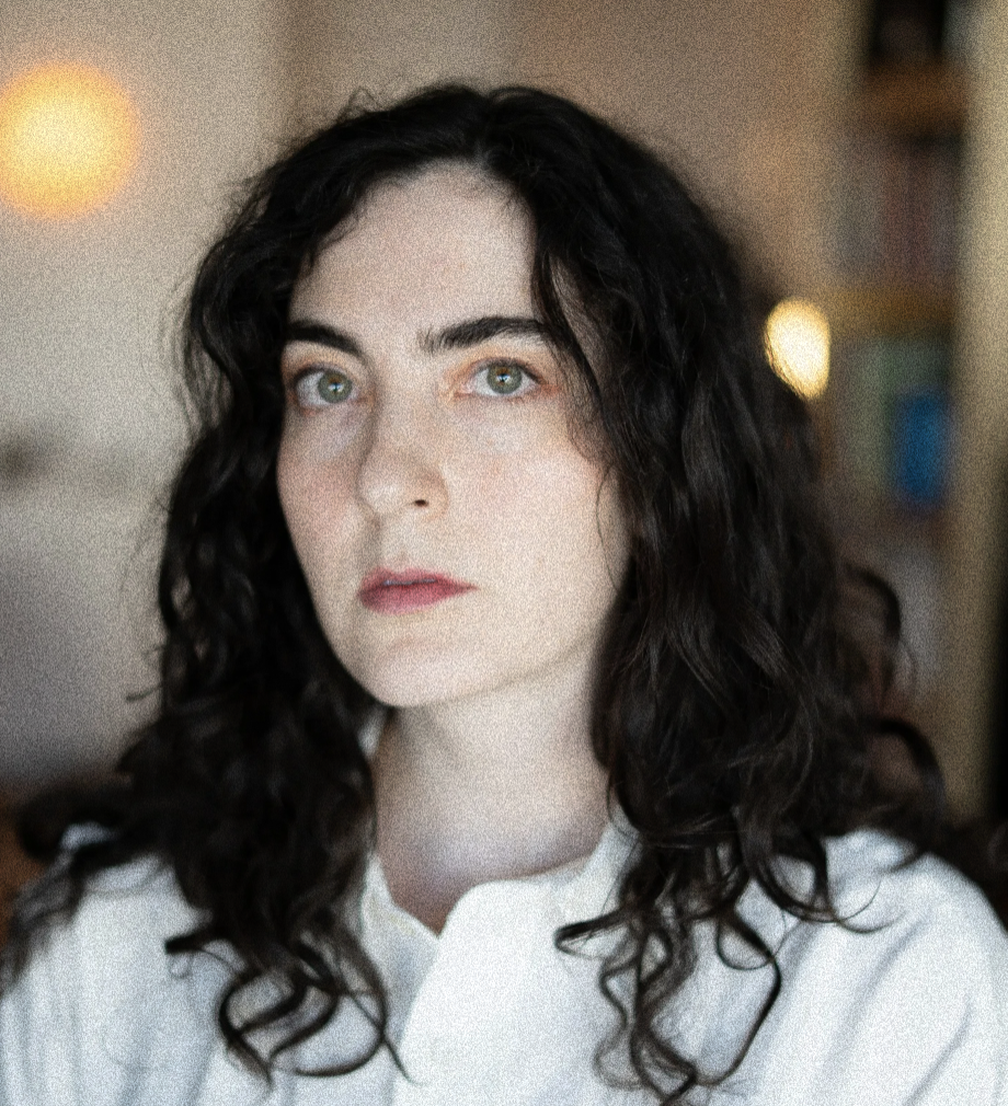 A white woman with black shoulder length, curly hair. She has green eyes and a white t-shirt. Her background is blurred out.