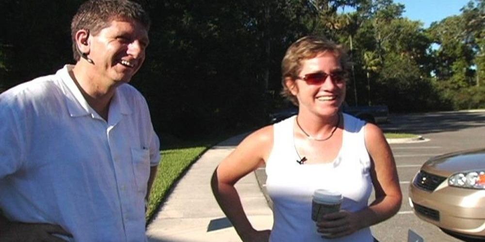 A man and a woman stand next to each other in a parking lot. They smile at something off camera.