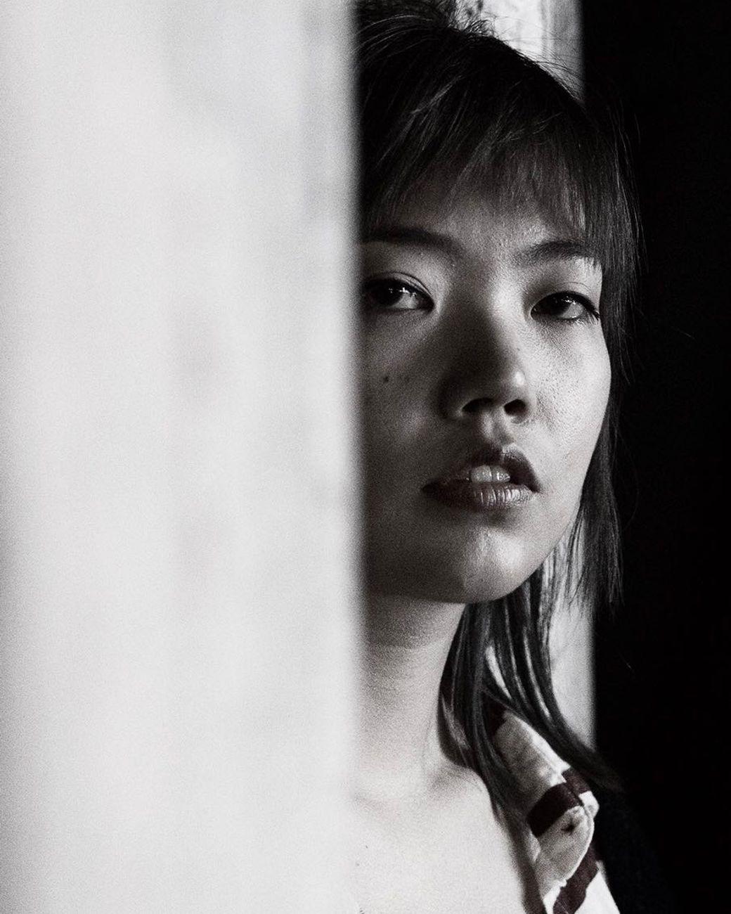 A black-and-white portrait of a woman looking into the camera, her face partly obscured by a wall.