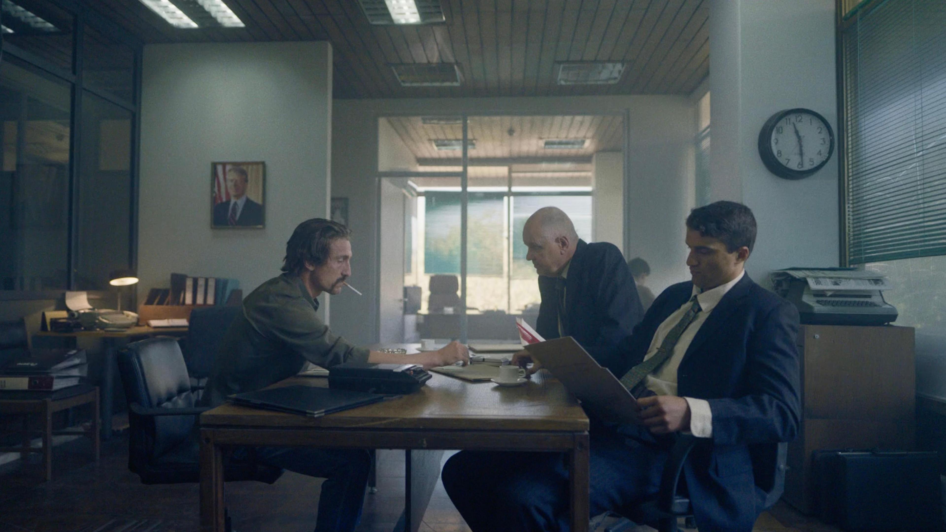 Three men in business attire sit around a table and look through documents.