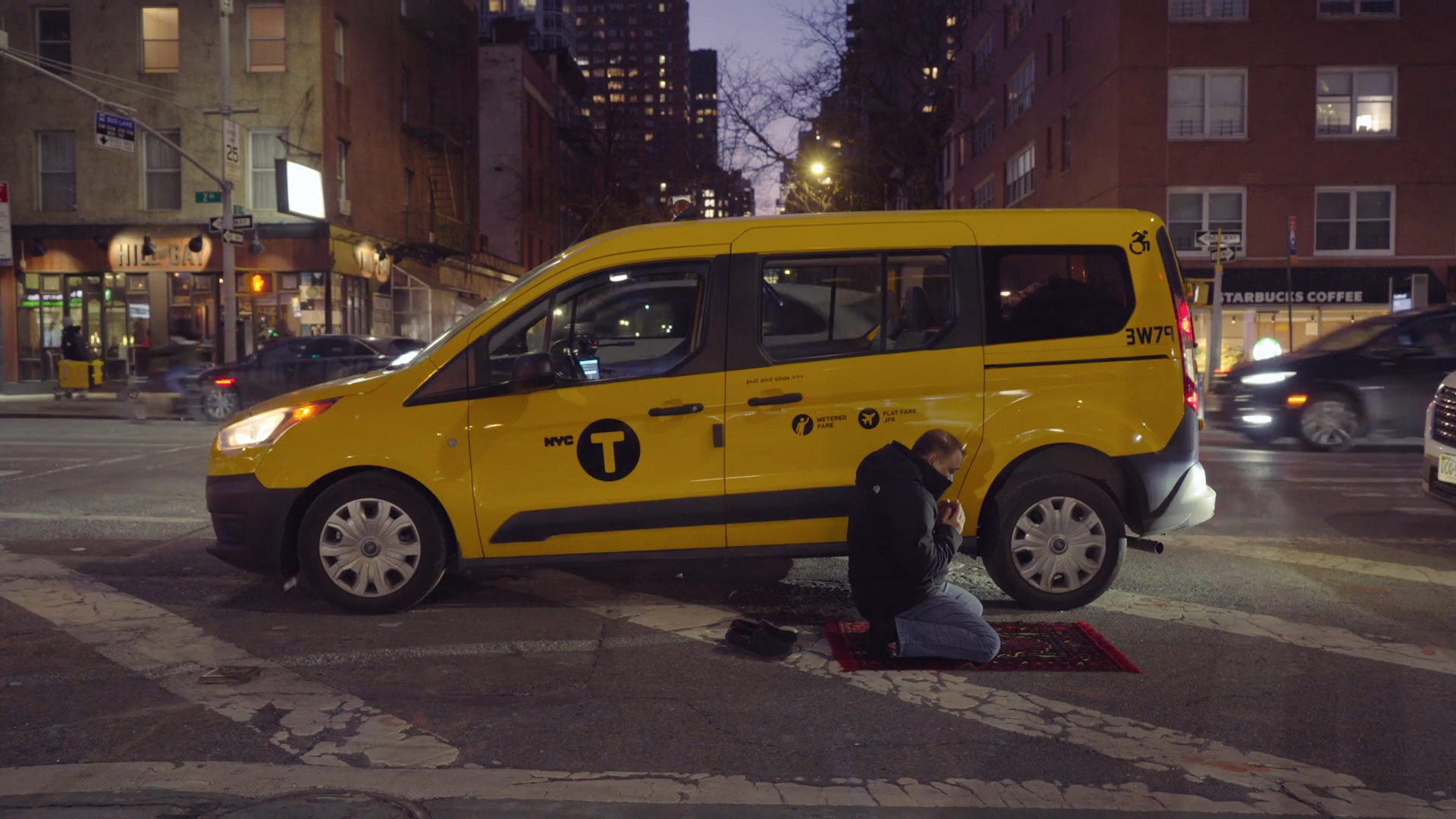 SK Mobinul Hoque, a New York Taxi Workers Alliance member and cab driver, kneels to pray on a prayer mat beside his yellow taxi at a New York City intersection in the evening.