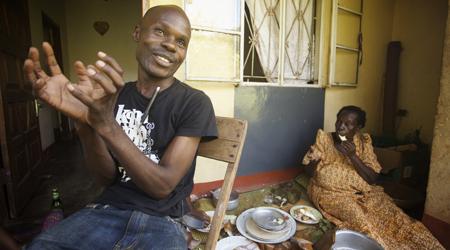 A man sits on a chair on a patio and speaks to the camera with a smile on his face. Near him is an elderly woman sitting amongst plates of food takes a bite of something.