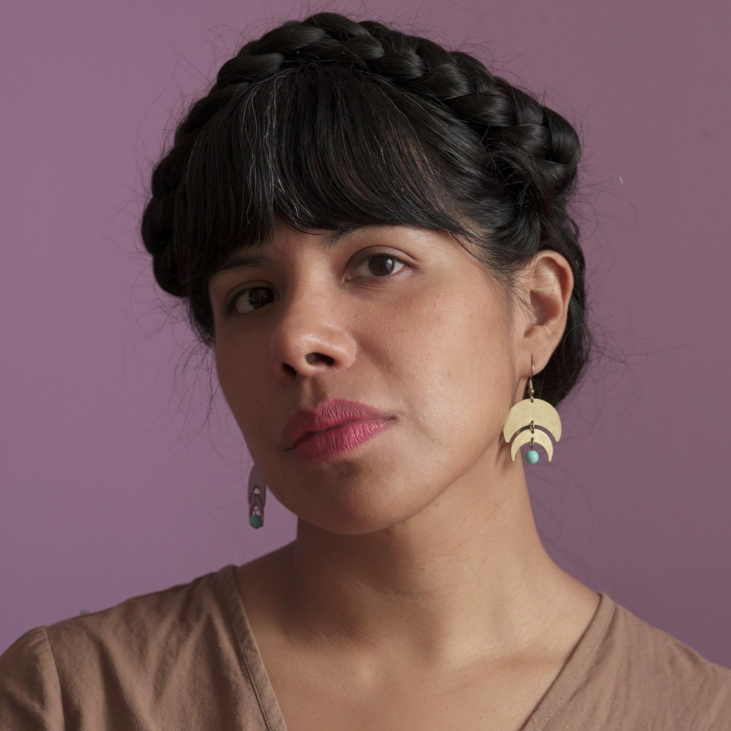 An editorial headshot featuring a Brown-skinned subject, with black braided hair arranged in a bun, photographed in soft natural light against a purple background. The person is wearing a brown blouse and golden earrings, with a thoughtful expression while their gaze meets the camera.