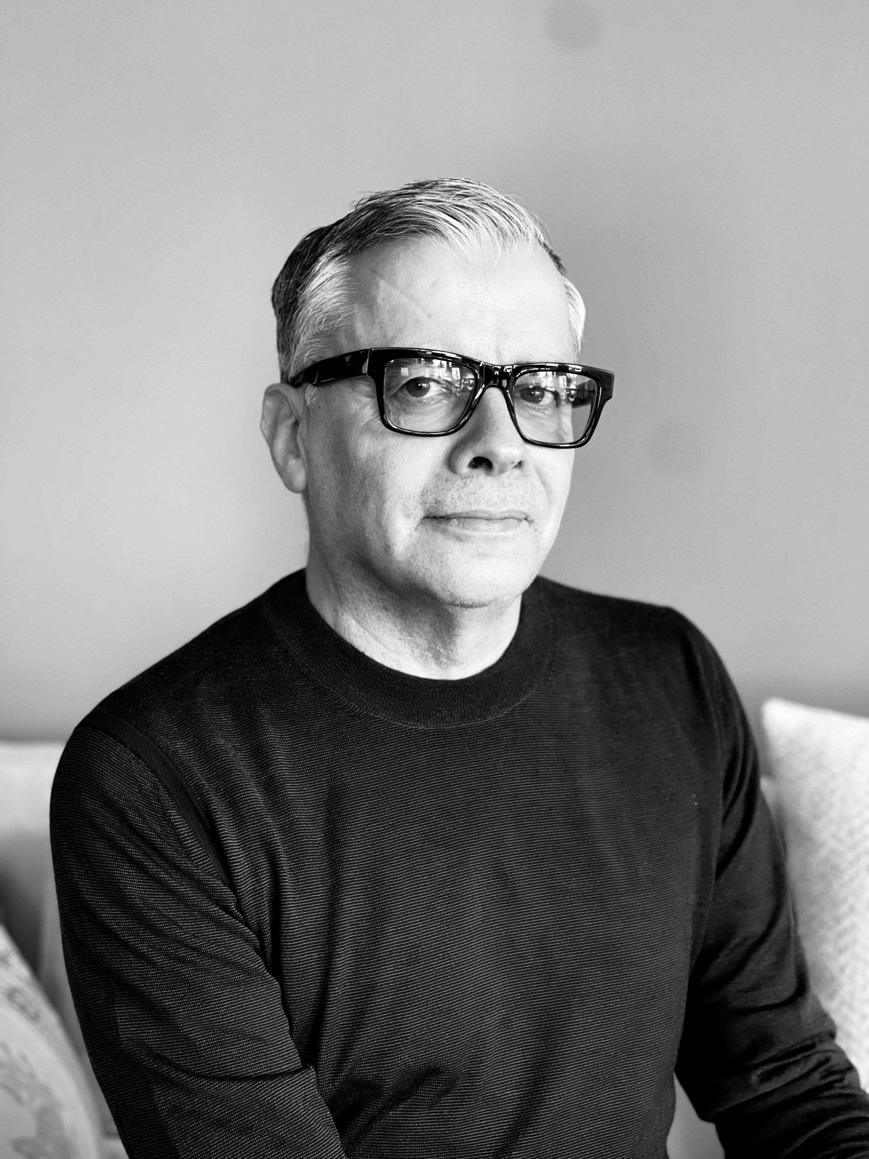 A black and white portrait of a man wearing glasses and a black shirt looking into the camera.