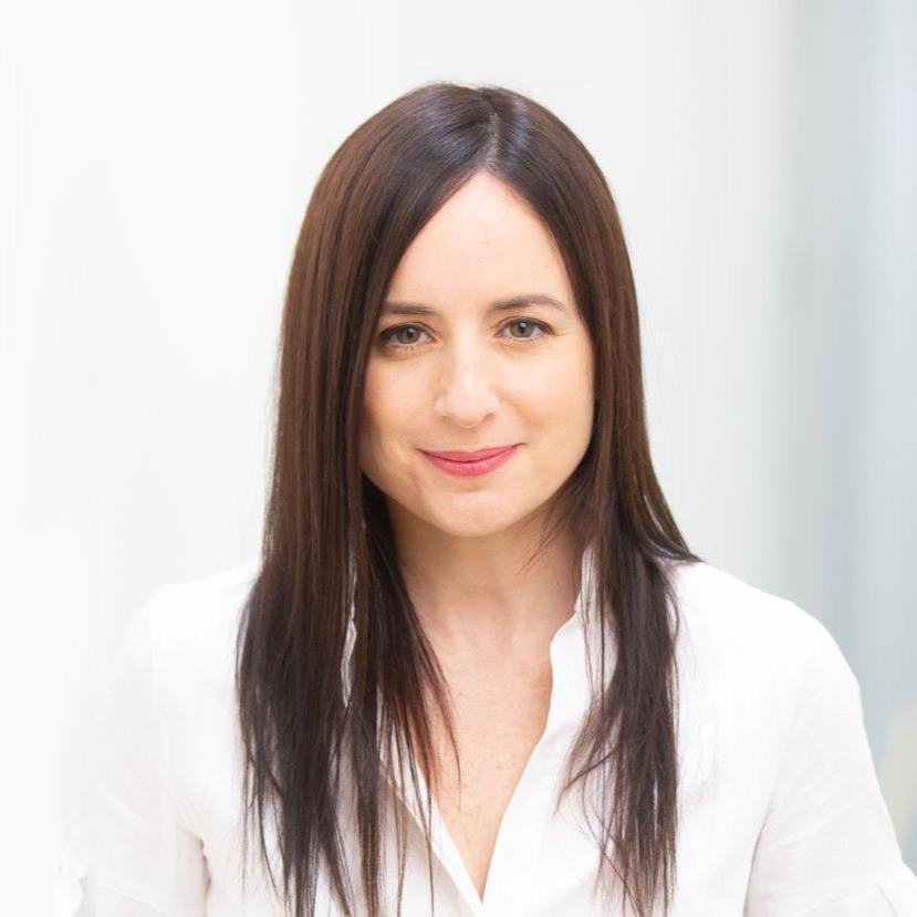 Portrait of filmmaker Maite Alberdi standing indoors, wearing a white blouse and blue jeans, looking at the camera with a slight smile against a softly blurred background.