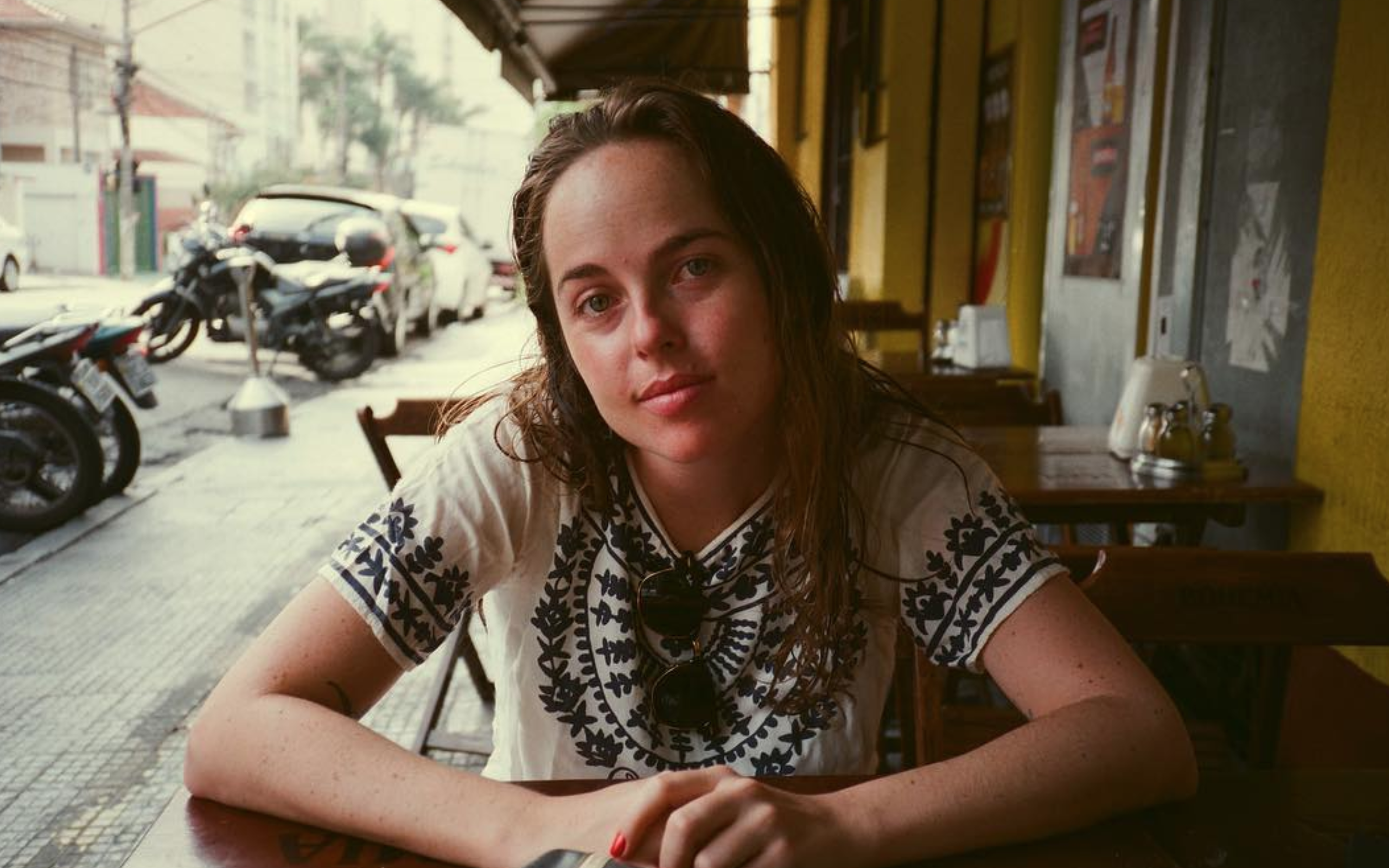Portrait of Sofia Geld sat and folding her hands in a table, wearing an embroidered shirt, and smiling slightly at the camera, she has medium long hair.