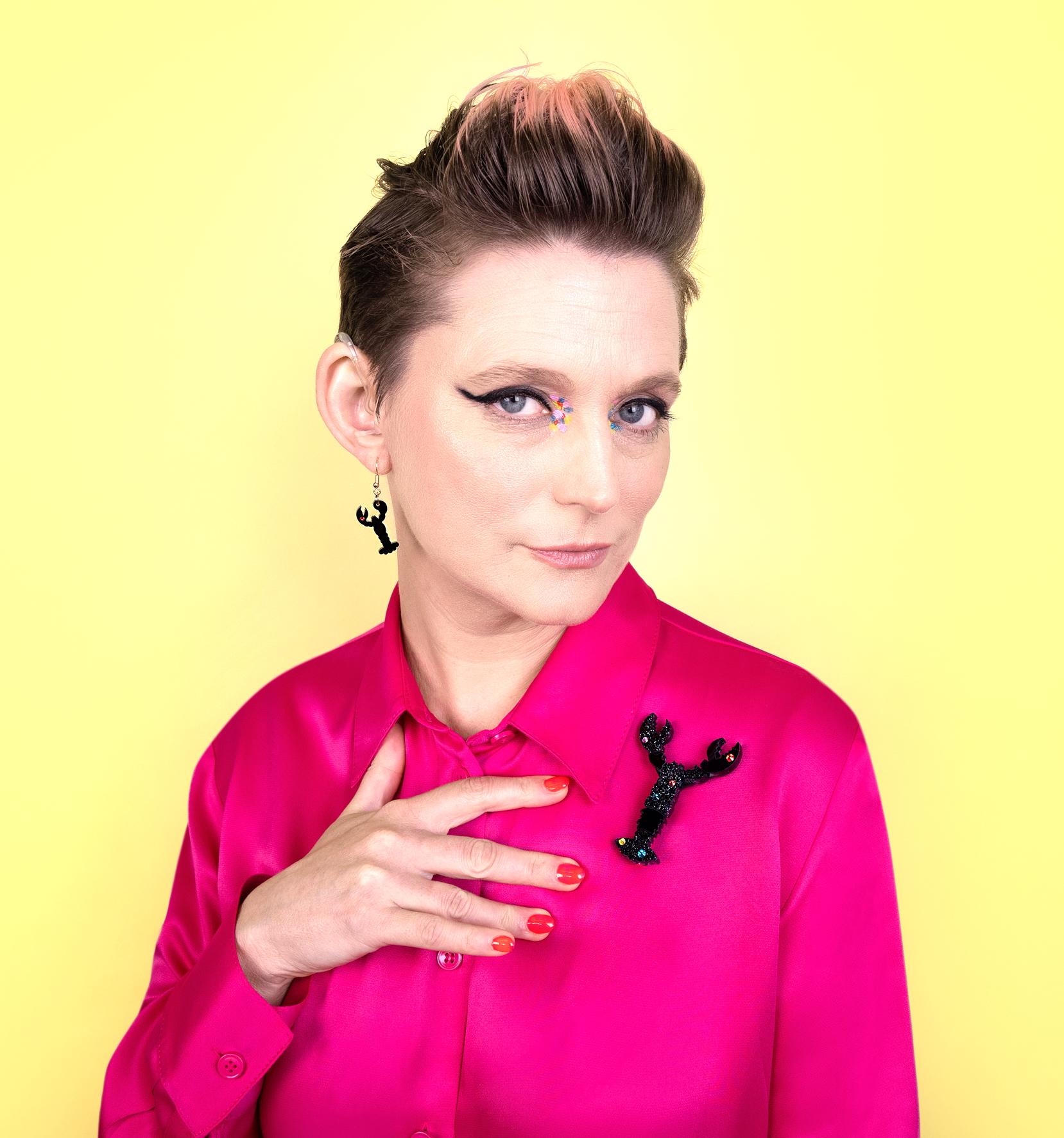 A 40-something white woman with pink hair and in a bright pink top. She is wearing hearing aids and has red finger nails.