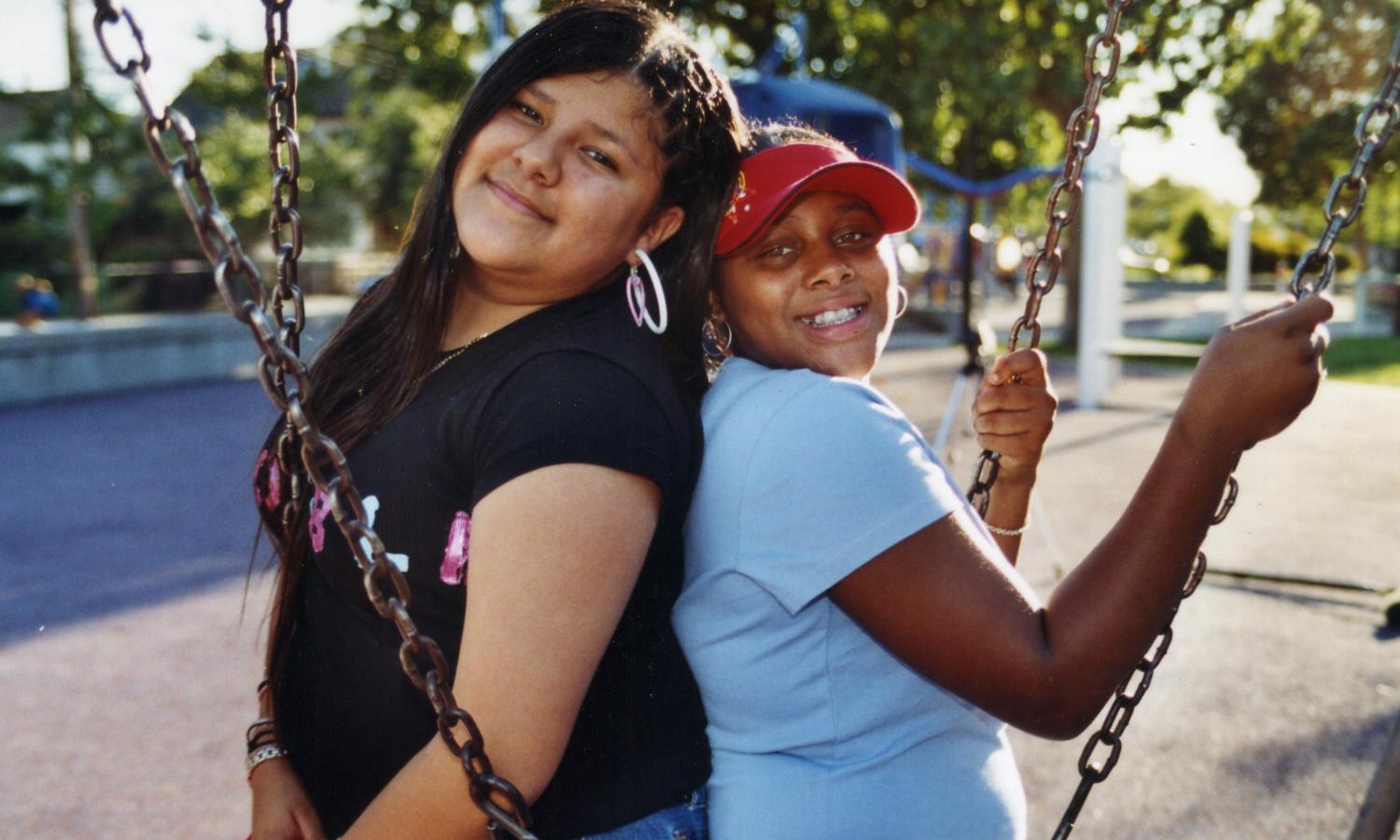 Two young girl on a playground stand with their backs against each other and smile into the camera. They hold onto the chains of swings.