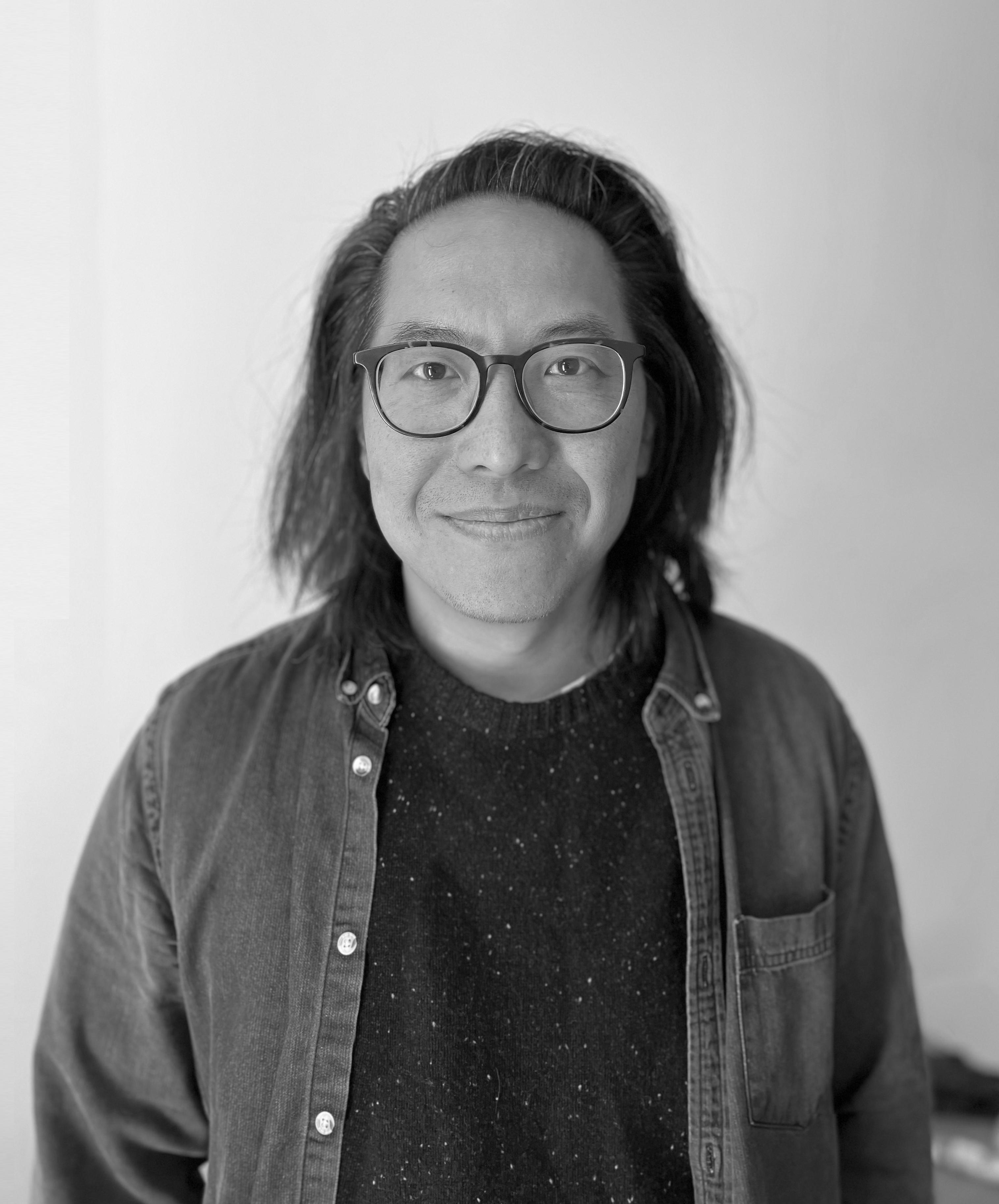 A black & white image of an Asian man with dark, shoulder-length hair wearing glasses and a dark shirt.