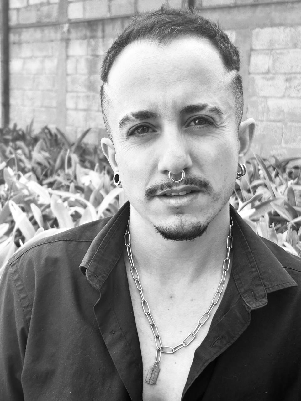Black-and-white headshot of a person with short dark hair, a mustache and goatee, wearing a septum ring, ear gauges, a silver chain necklace, and a dark collared shirt. They look directly at the camera against a softly blurred background of plants and a stone wall.