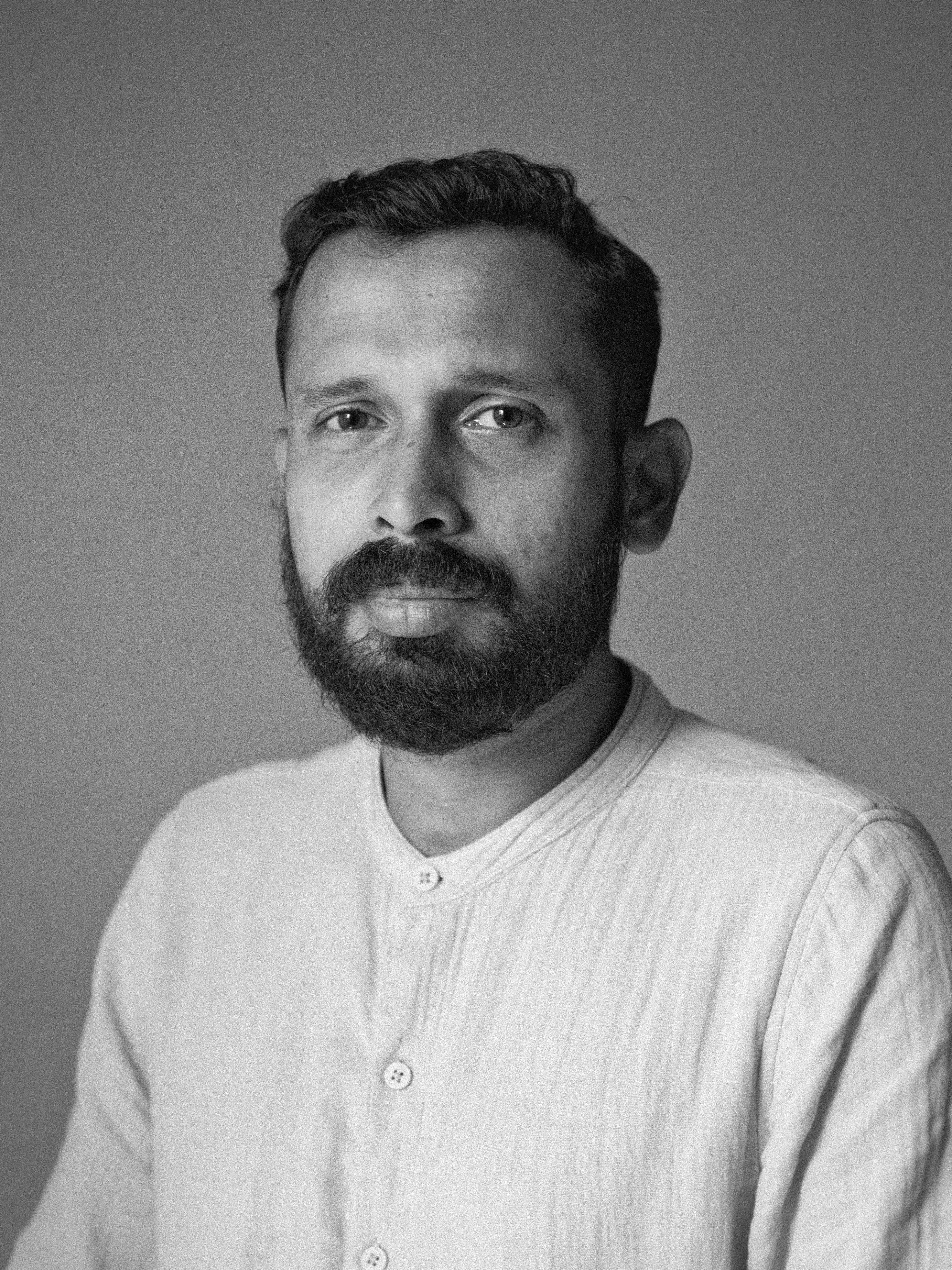 Black and white portrait of man sporting a beard and a button up light shirt looking at the camera slightly smiling.