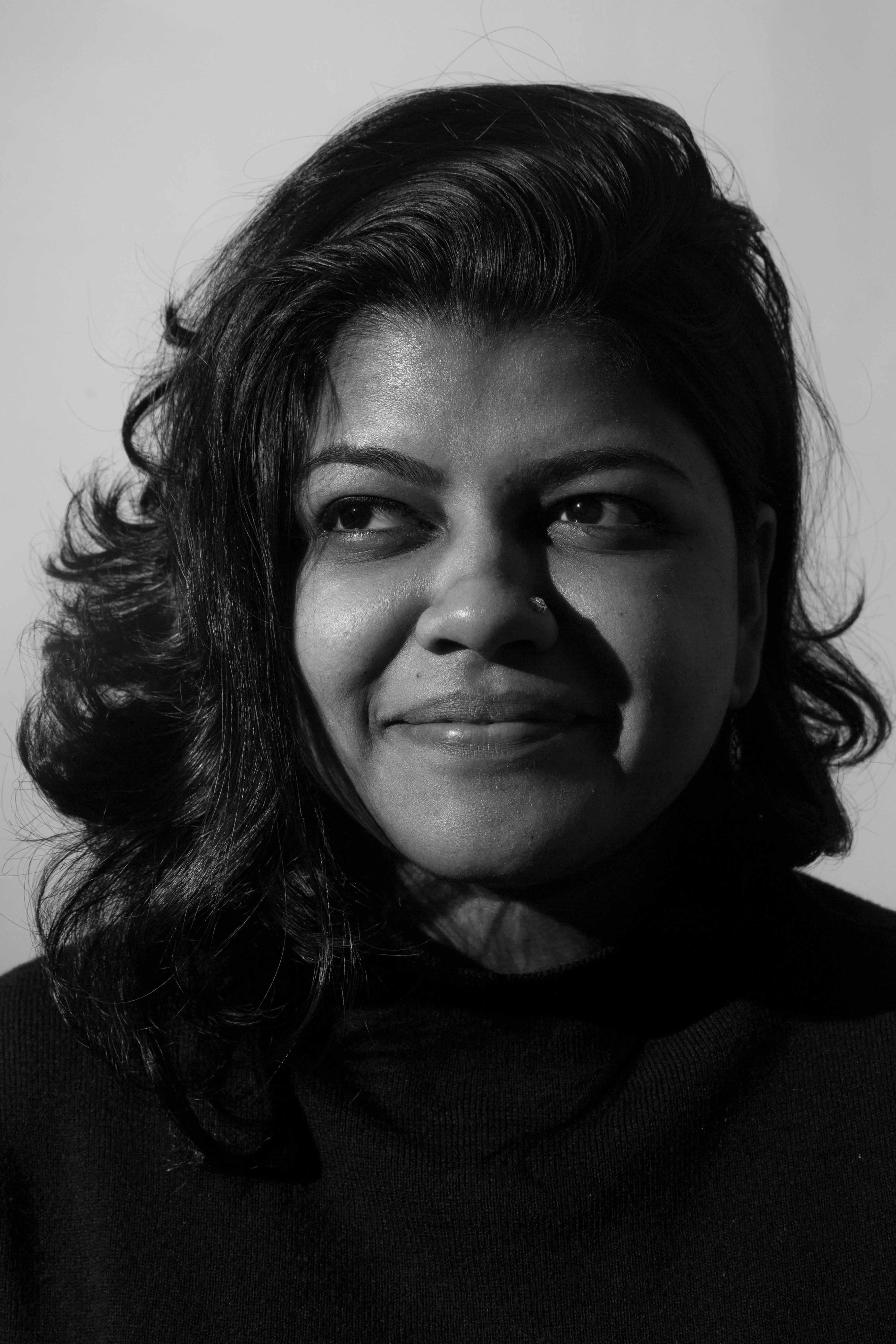 Headshot of an Indian woman with shoulder-length black hair, wearing a black shirt and smiling sideways against a neutral gray background.