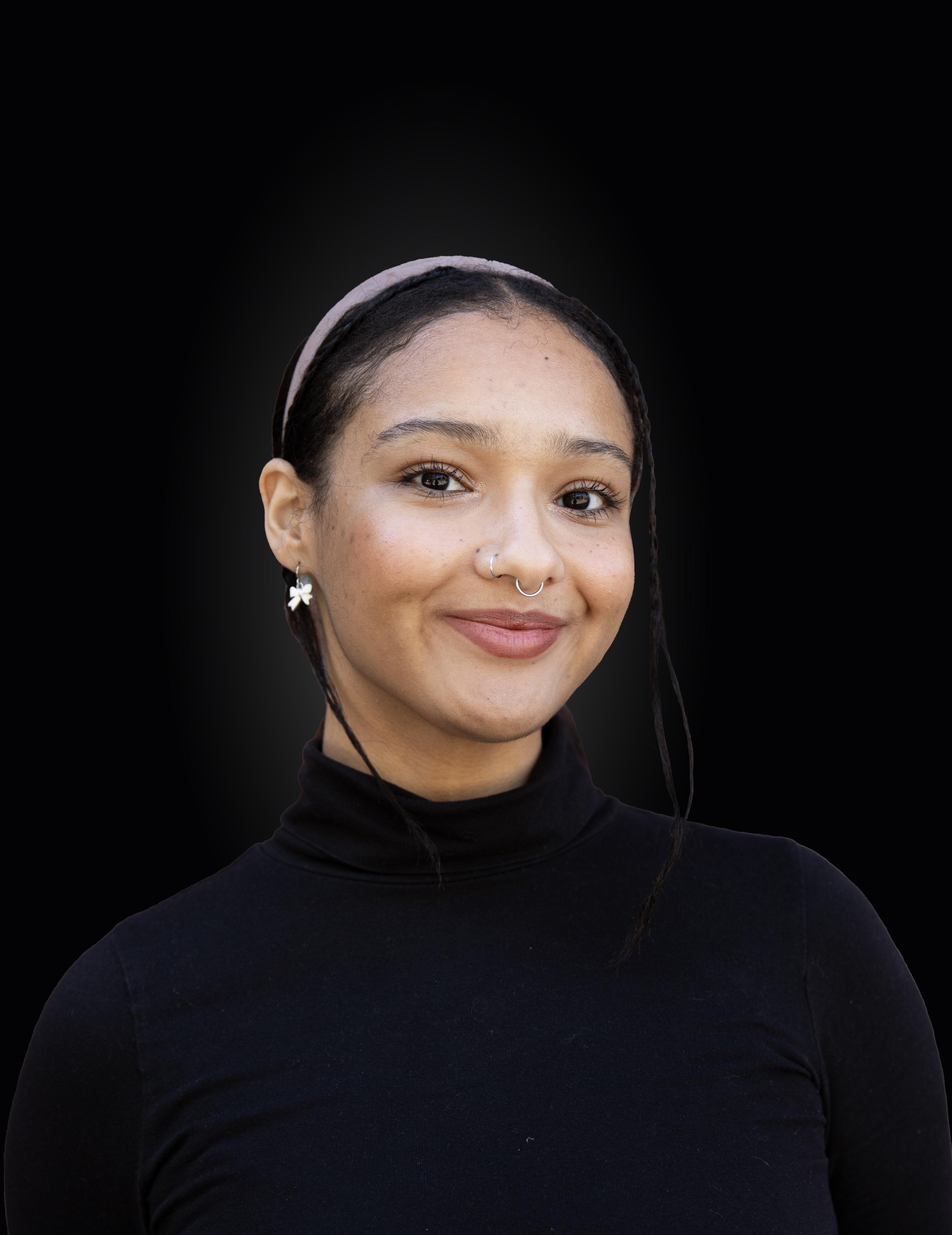A woman stands in front of a dark backdrop and smiles into the camera. She is wearing a black turtleneck and a baby pink headband. Her hair is pulled back with strands of braids framing her face.