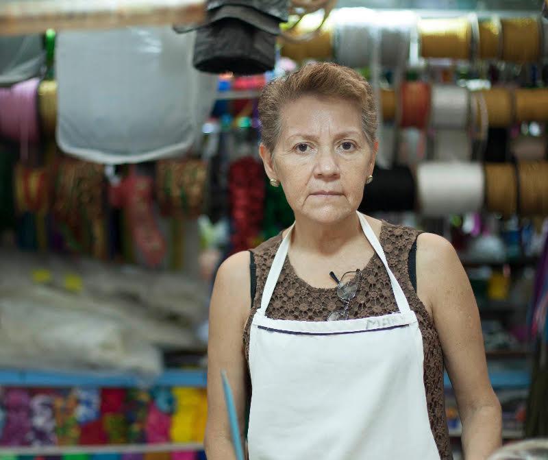An elderly woman wears an apron and stands in front of a fabric display.