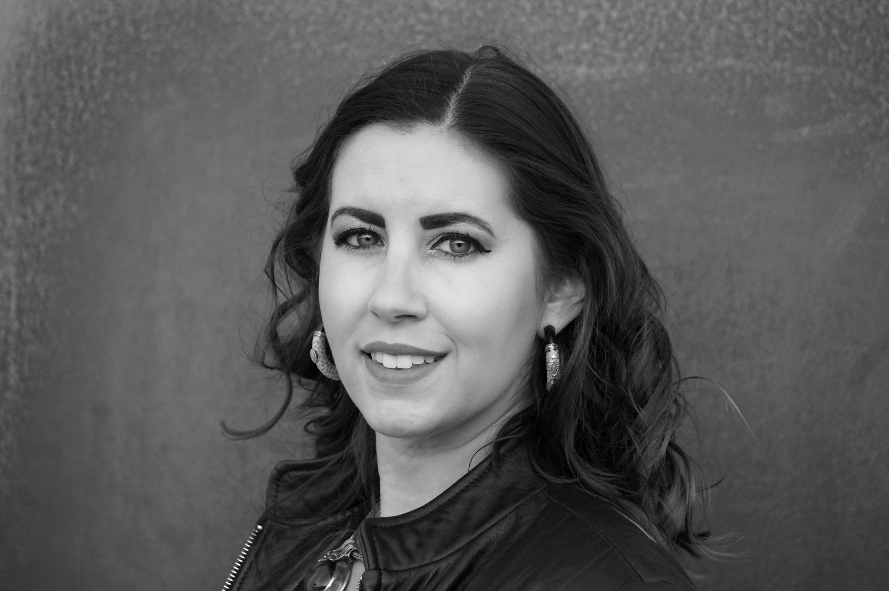 black and white headshot of filmmaker Erika Cohn. a young woman with shoulder length, dark wavy hair looks directly at the camera. She has light colored eyes ringed with dark eyeliner and wears large silver earrings.