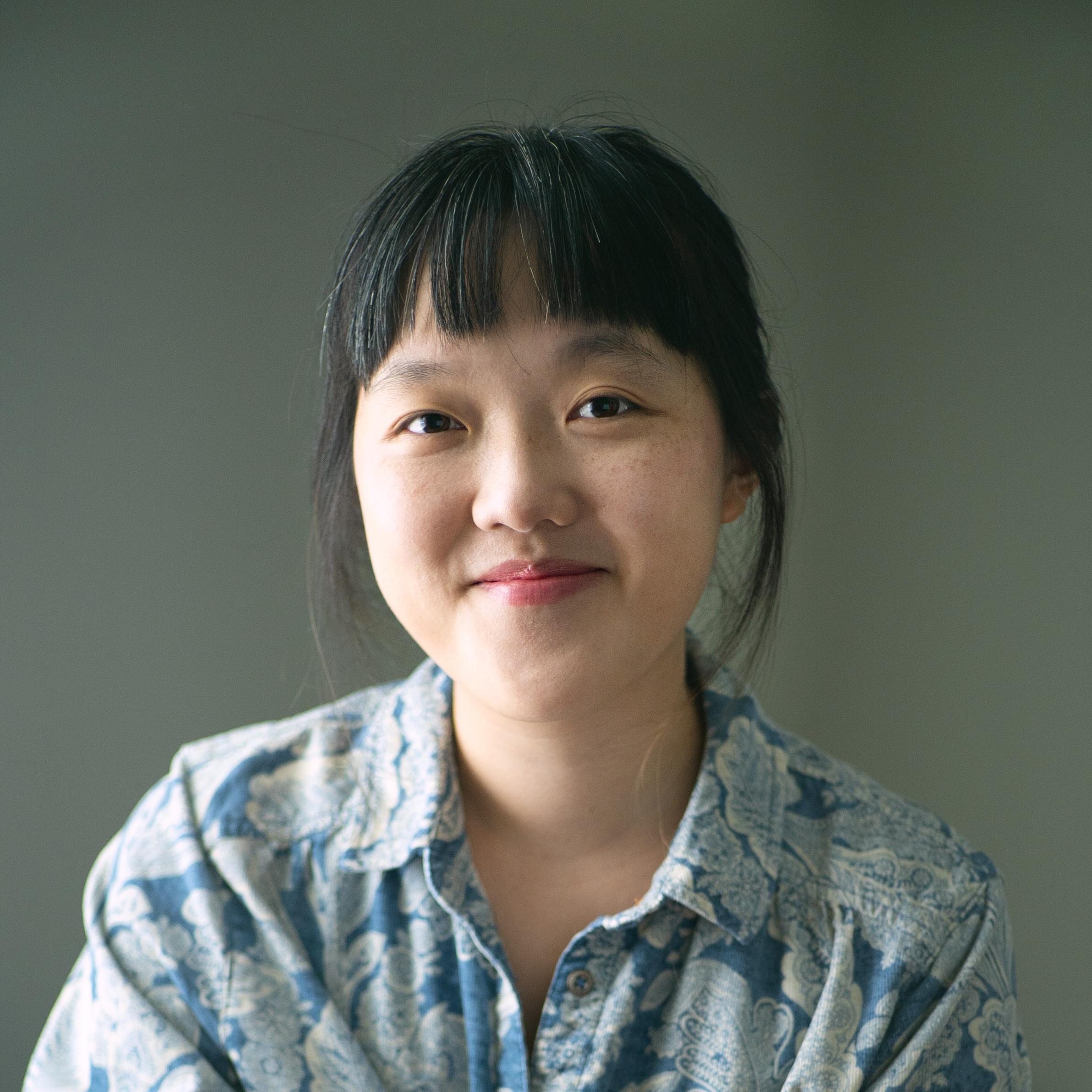 A woman sits in front of a sage background and smiles at the camera. She has dark bangs and is wearing a blue button down shirt
