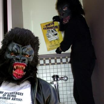 Two people dressed in gorilla costumes stand in a bathroom stall. One of them puts a poster on the wall that reads "The Birth of Feminism."