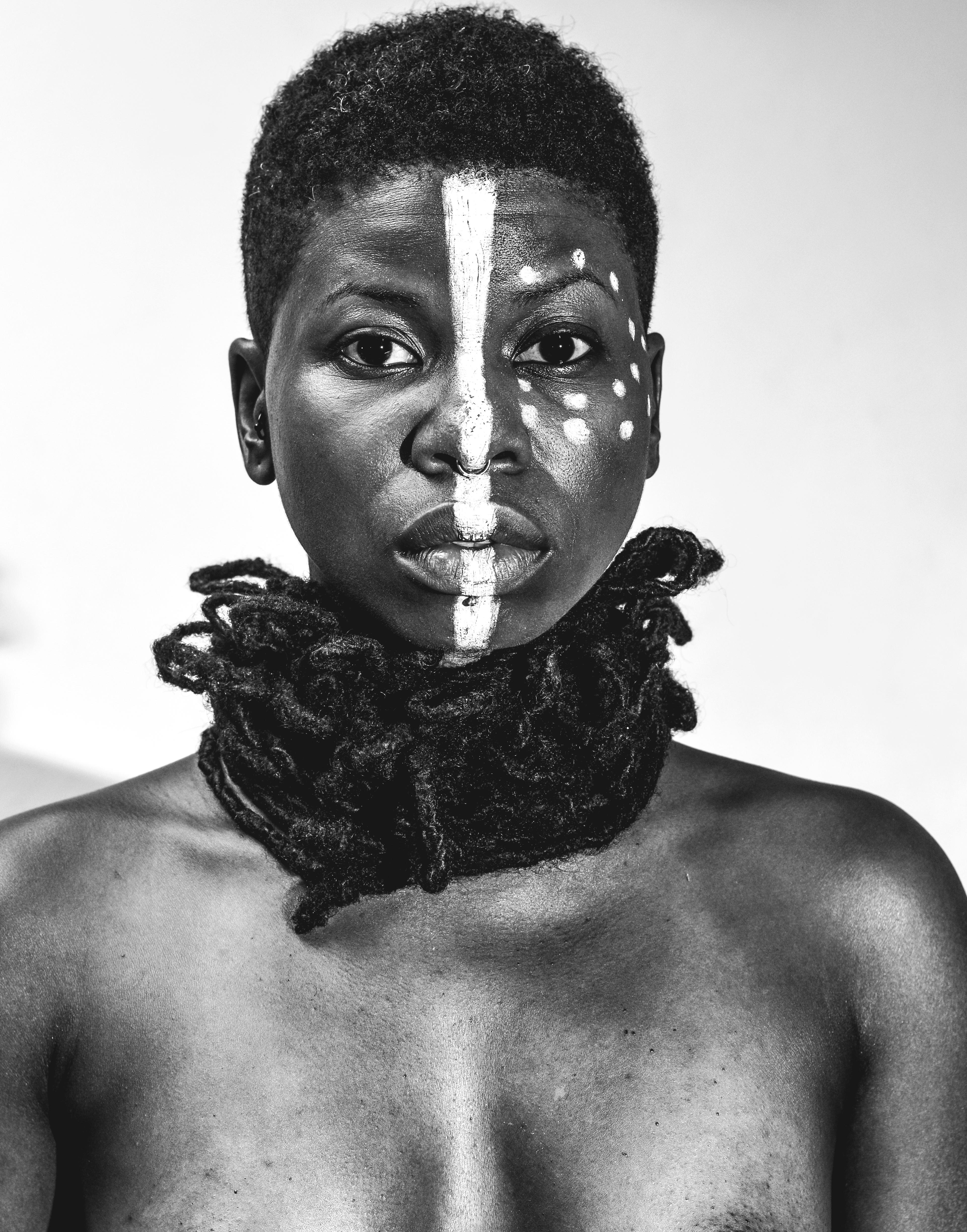 A black and white portrait a person with white paint splitting their face down the middle and dots circling their right eye.