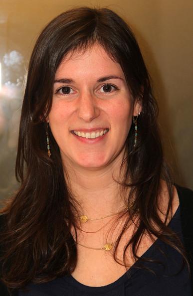 Madeline Sackler looks directly at the camera and smiles. She has long brown hair and is wearing two earings and a gold necklace.