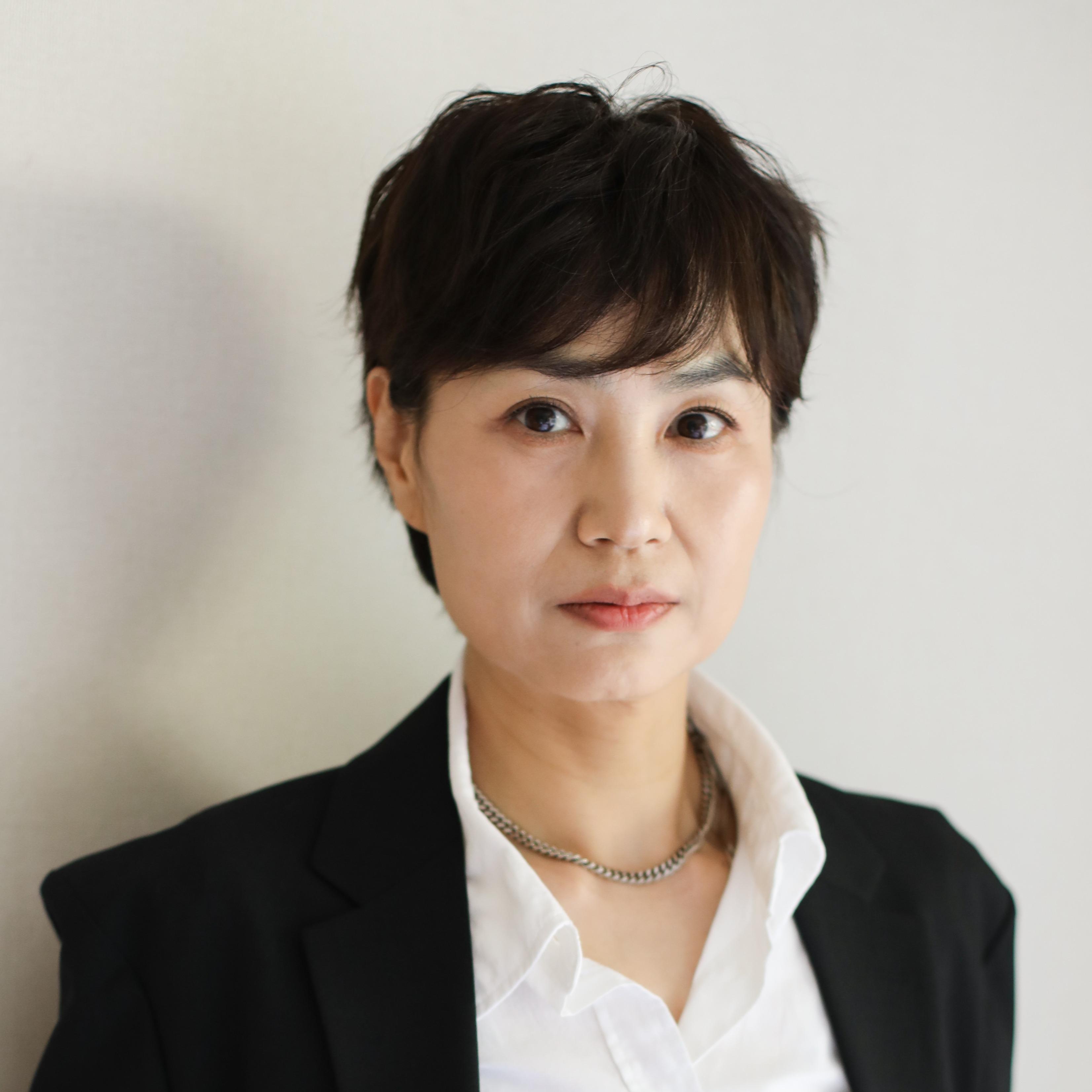 Ilrhan with short dark hair stands against a plain light wall, wearing a black blazer and white shirt, looking calmly at the camera.