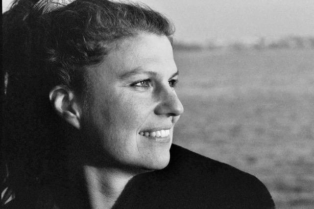 Katja Esson standing in profile to the camera. She is looking off camera to her right and is wearing a coat. Background is out of focus. Black and white portrait.