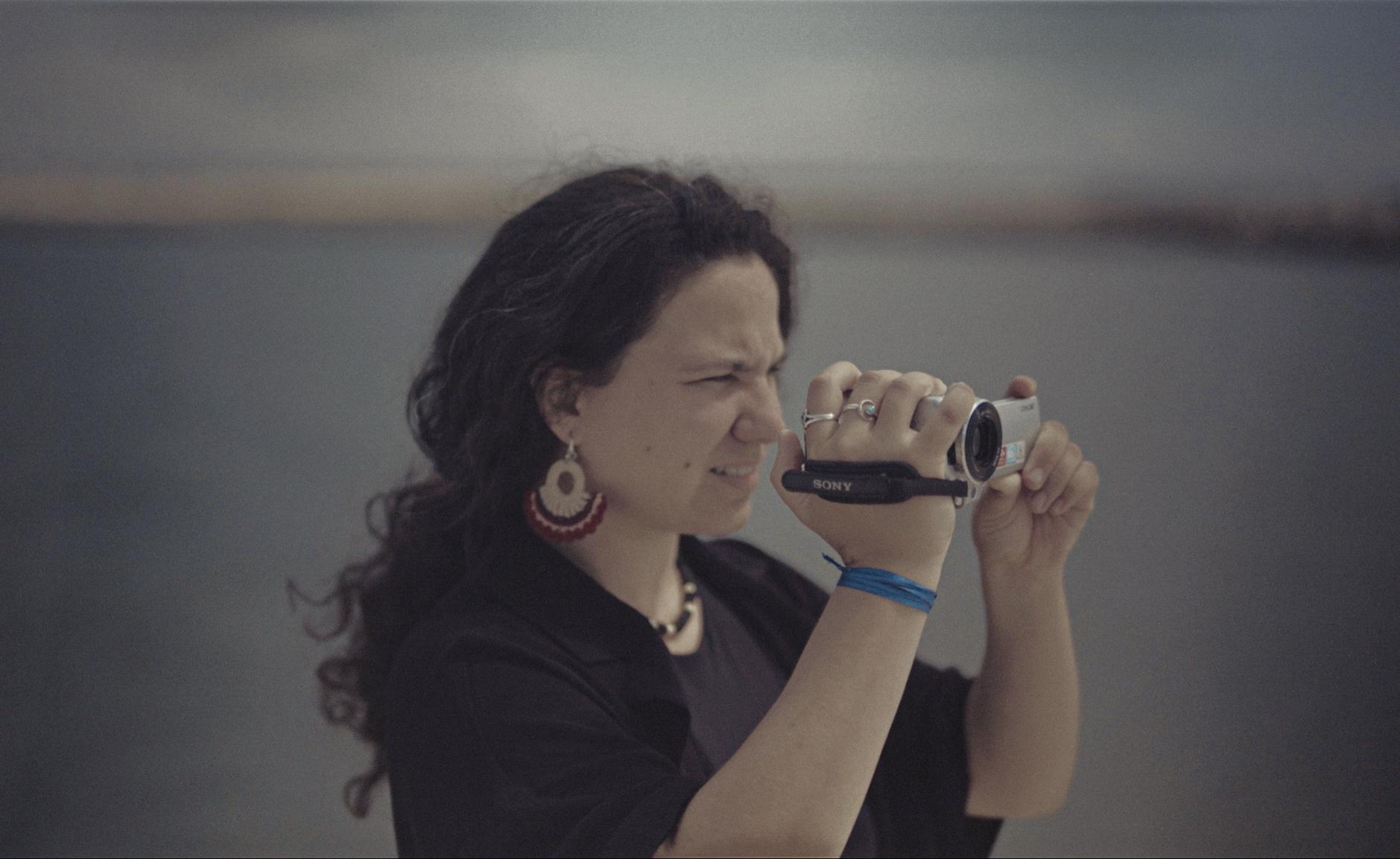 Color photograph of a young brunette woman filming with a handycam, with the sea out of focus in the background.